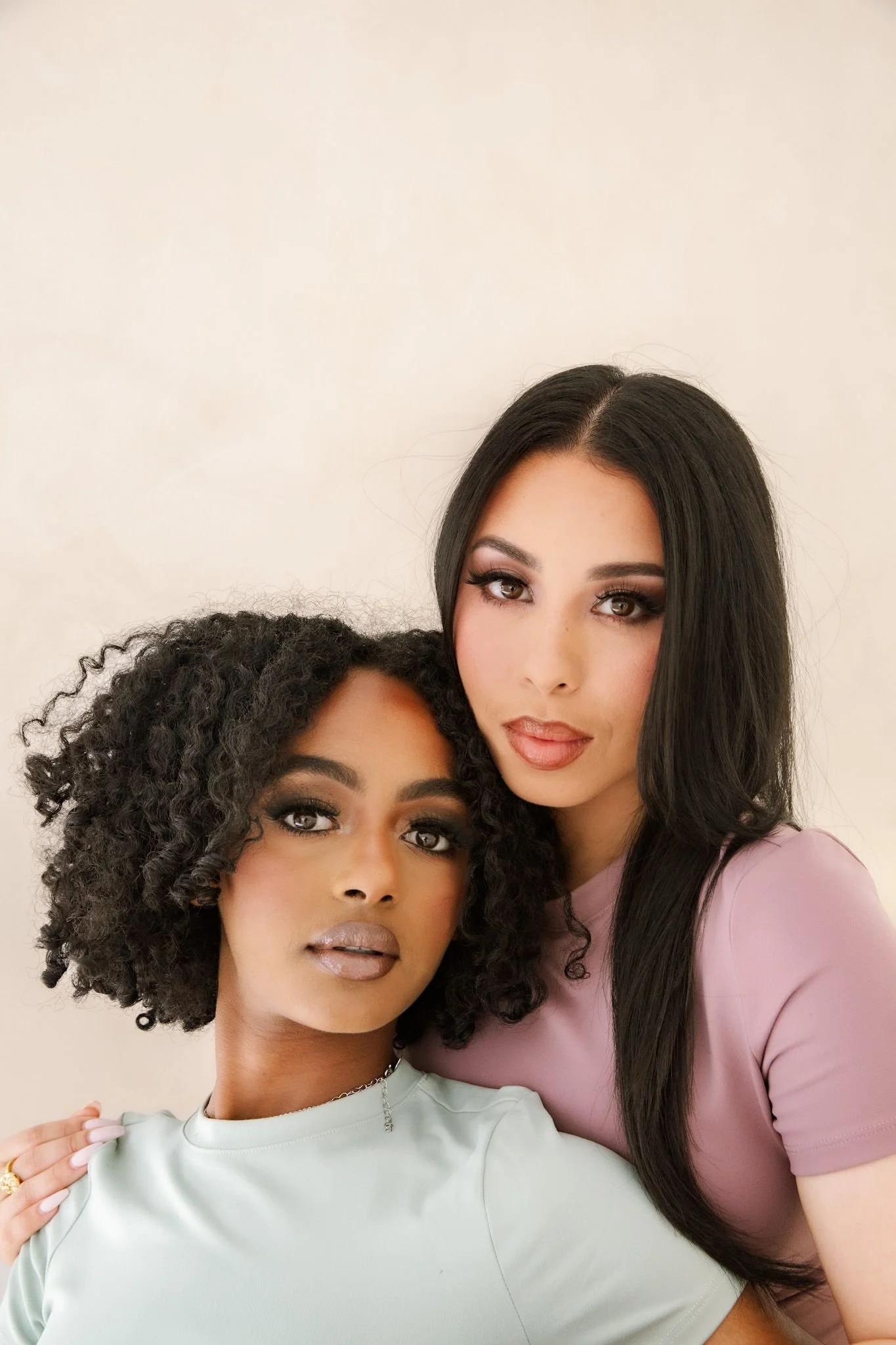 Two women, one with curly hair and the other with straight, long dark hair, posing closely together against a neutral background.