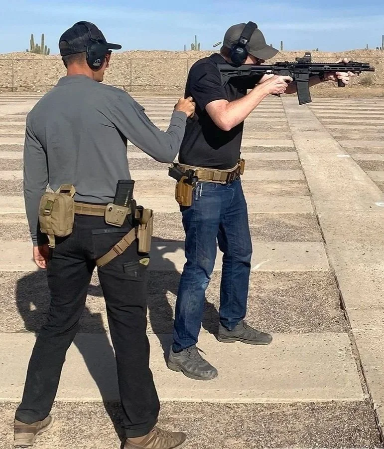 Two men at a shooting range, one aiming a rifle while the other assists by holding his arm. Both are wearing hats, ear protection, and have gun gear.