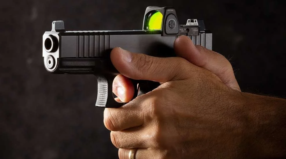 A person holding a handgun with a green reflex sight mounted on it.
