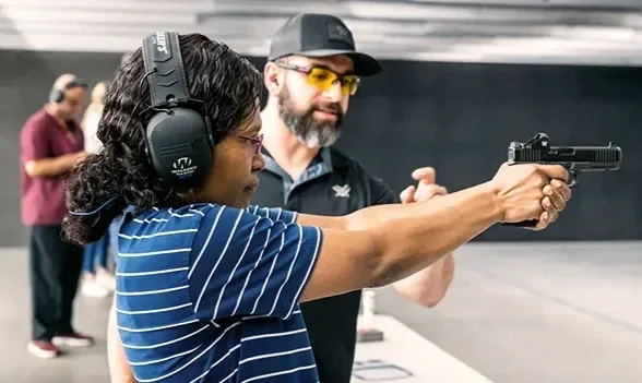 Woman at indoor shooting range holding a pistol with instructor guiding her.