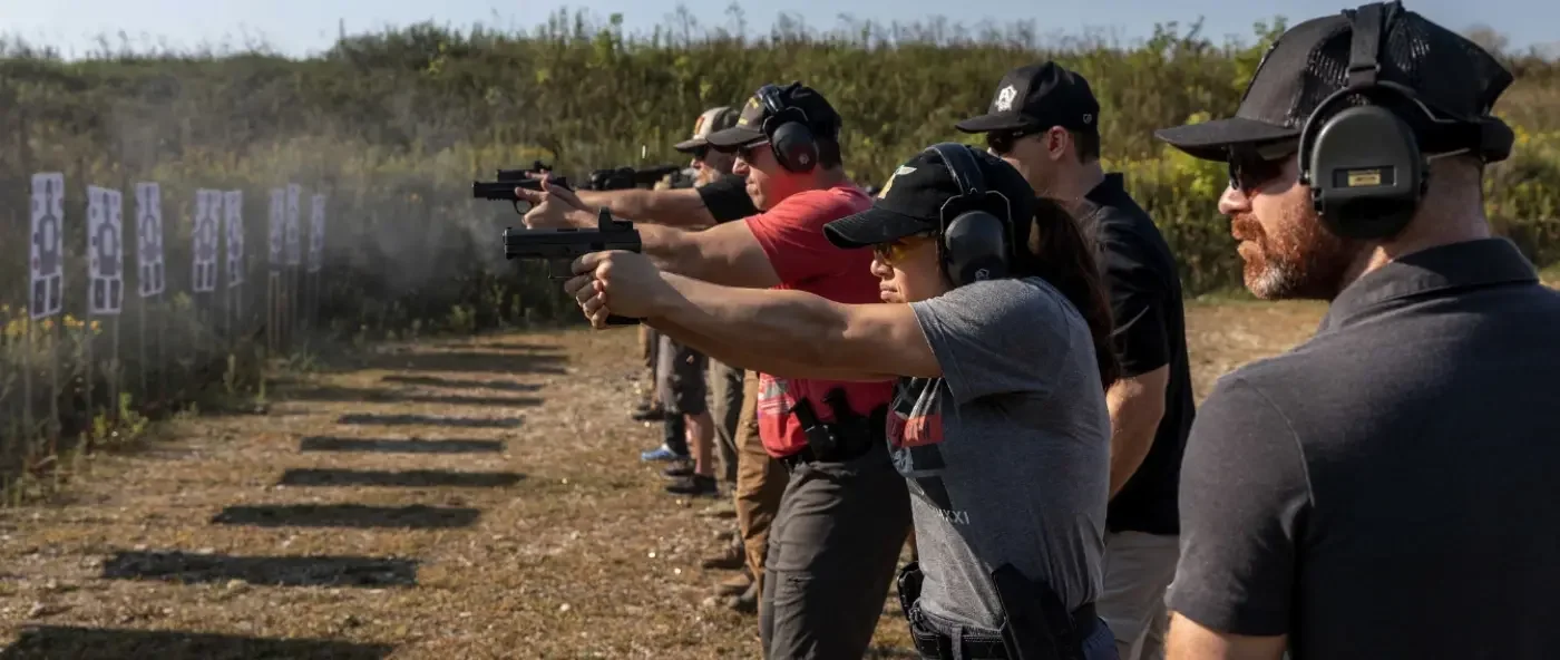 A group of people at an outdoor shooting range, aiming pistols at targets, wearing ear protection and sunglasses.