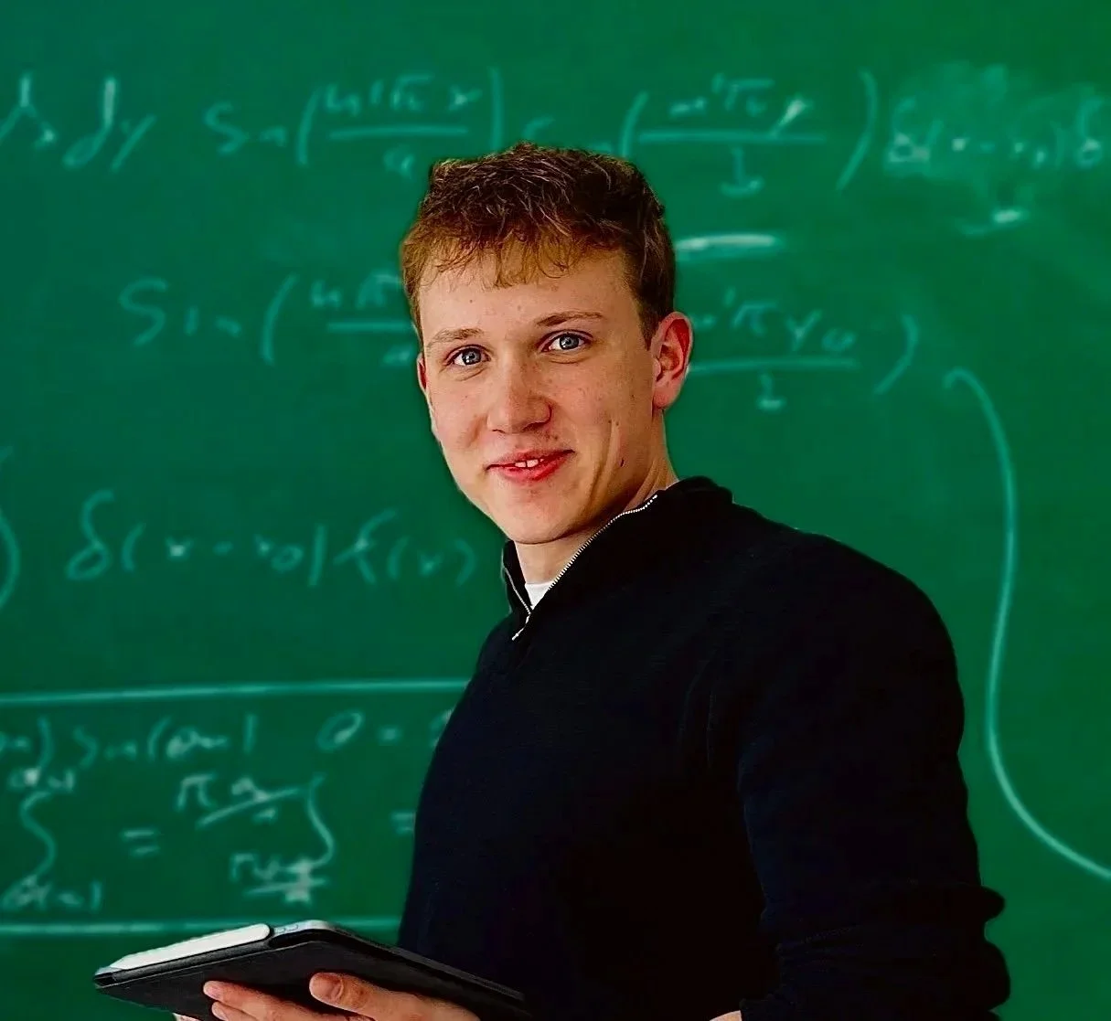 A young man with short brown hair and blue eyes standing in front of a green chalkboard filled with mathematical equations, holding a tablet in his hand and looking at the camera with a slight smile.