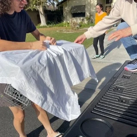 The Whisker Project of Kern-TNR:  People gathered around a grill covered with a white cloth outdoors, with one person sitting at the table and others standing nearby.