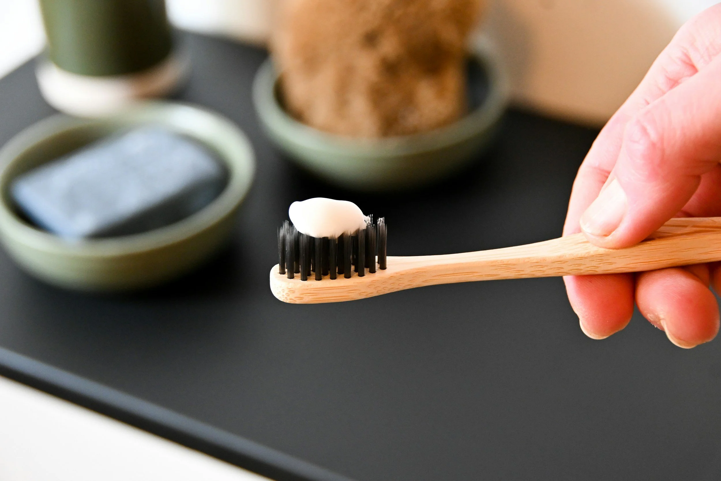 A close-up of a hand holding a wooden toothbrush with white facial cream on black rubber bristles, with green bowls containing soap and a mineral stone in the background.