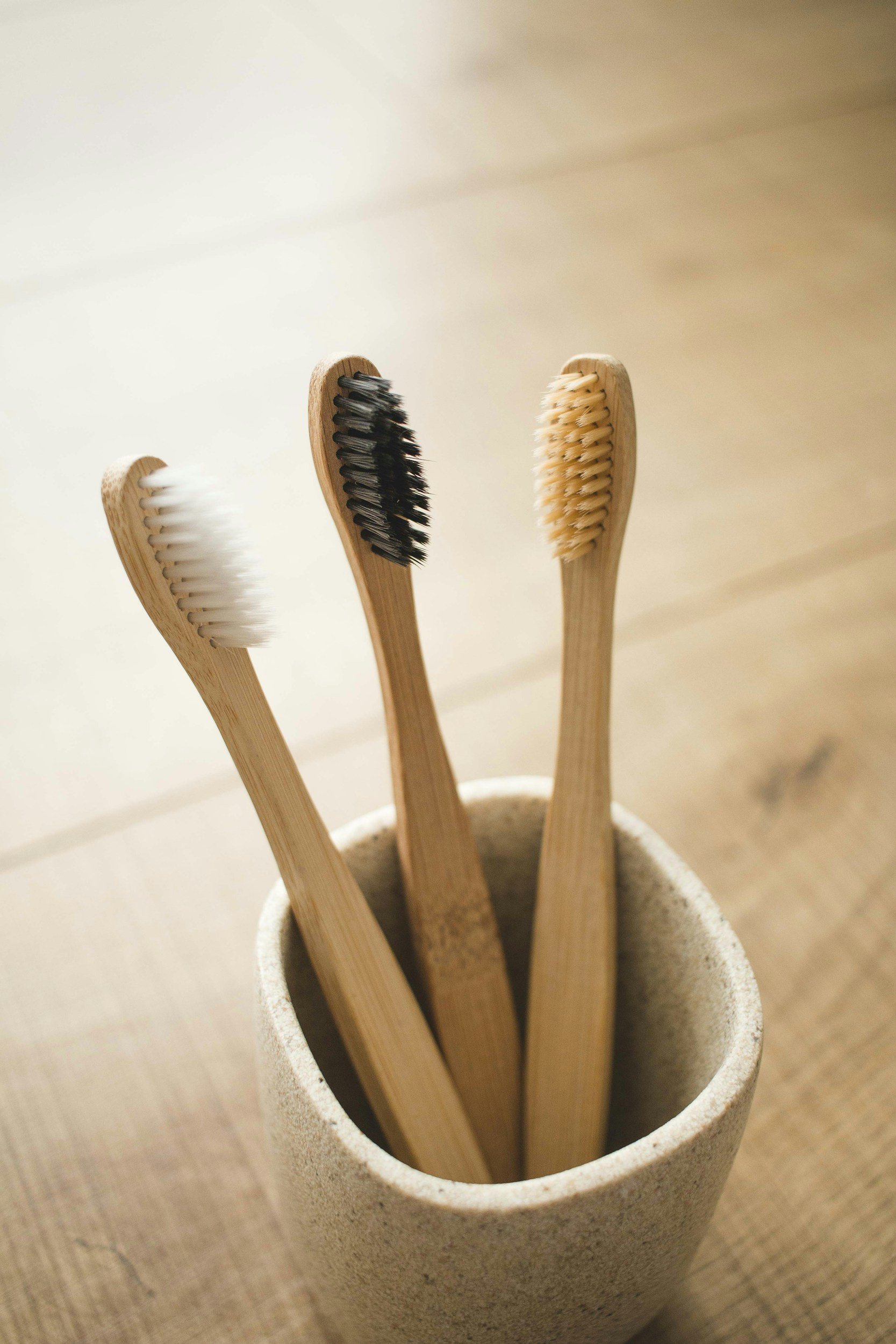 Three bamboo toothbrushes with different colored bristles in a beige ceramic holder on a wooden surface.