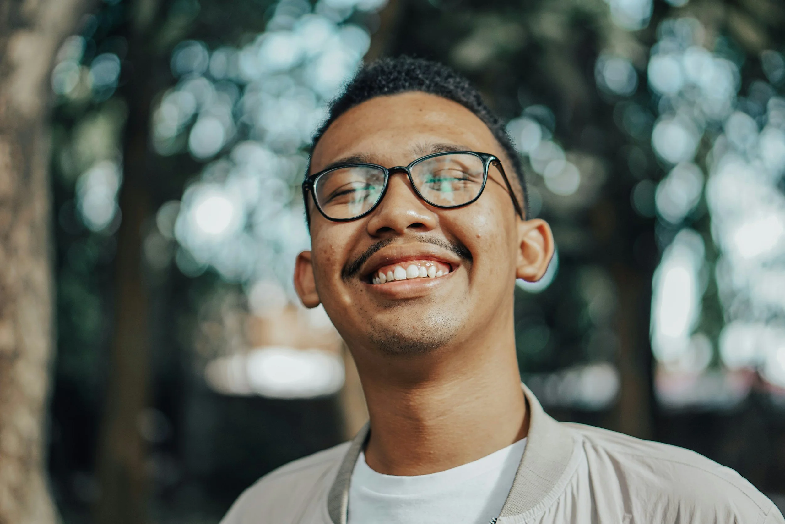 Smiling young man with glasses outdoors with trees and bokeh background