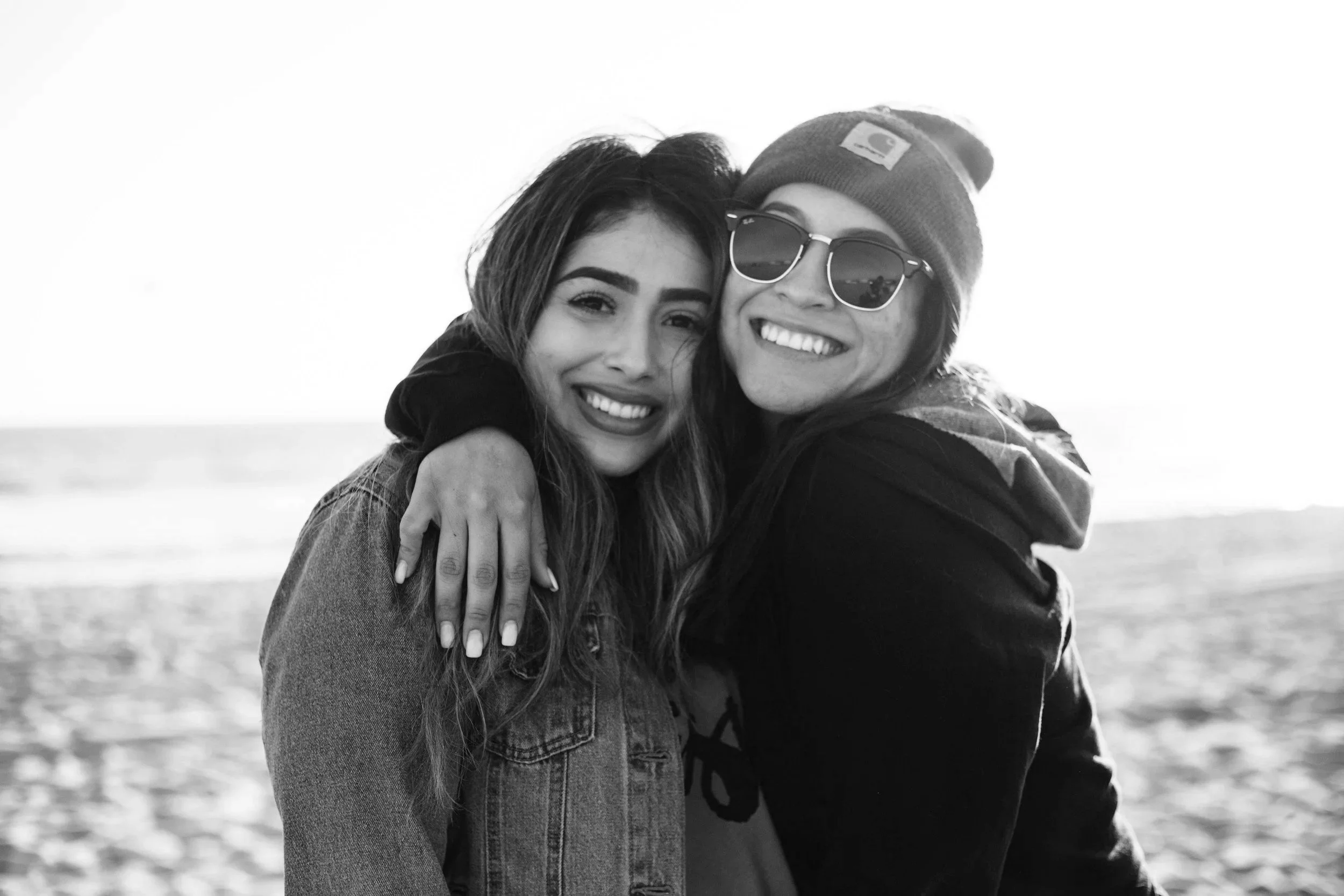 Two young women hugging and smiling on the beach, one wearing sunglasses and a beanie, the other with long hair and a denim jacket, black and white photo.