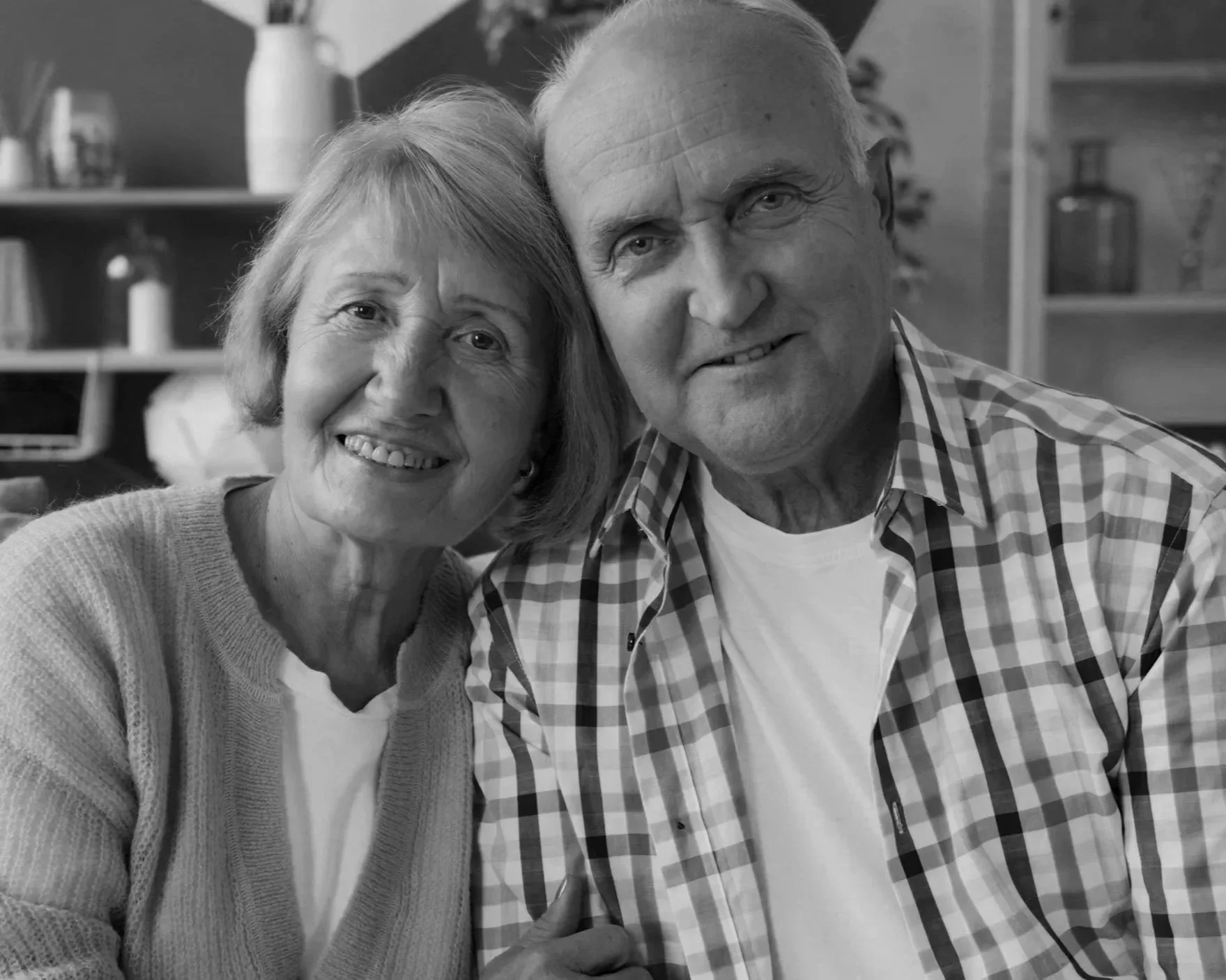 A smiling elderly woman and elderly man with their heads leaning together in a cozy kitchen setting.