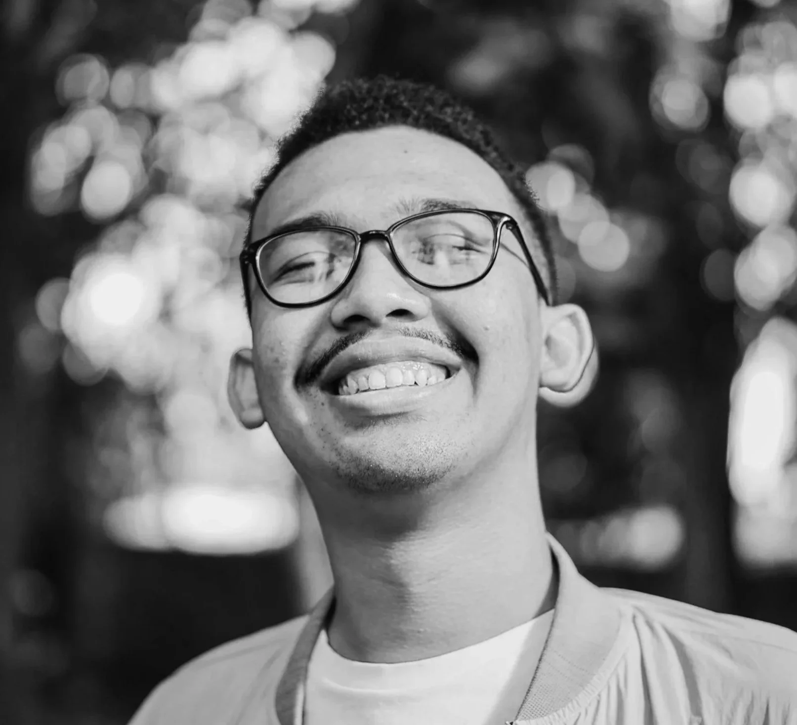 Black and white photo of a smiling man with glasses, short hair, and facial hair, outdoors with blurred trees and bokeh lights in the background.