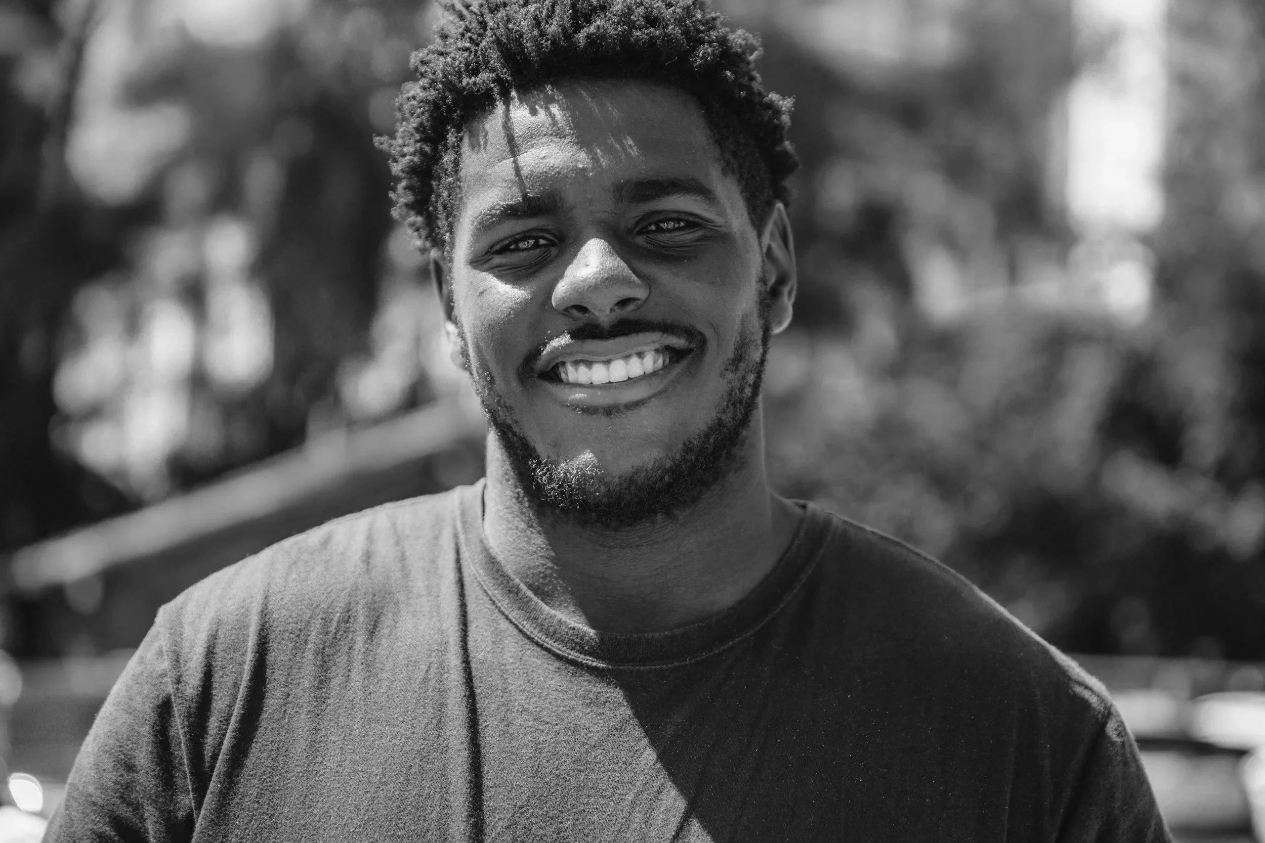 A smiling young man with curly hair and a beard, wearing a t-shirt, standing outdoors with blurred trees and sunlight in the background.