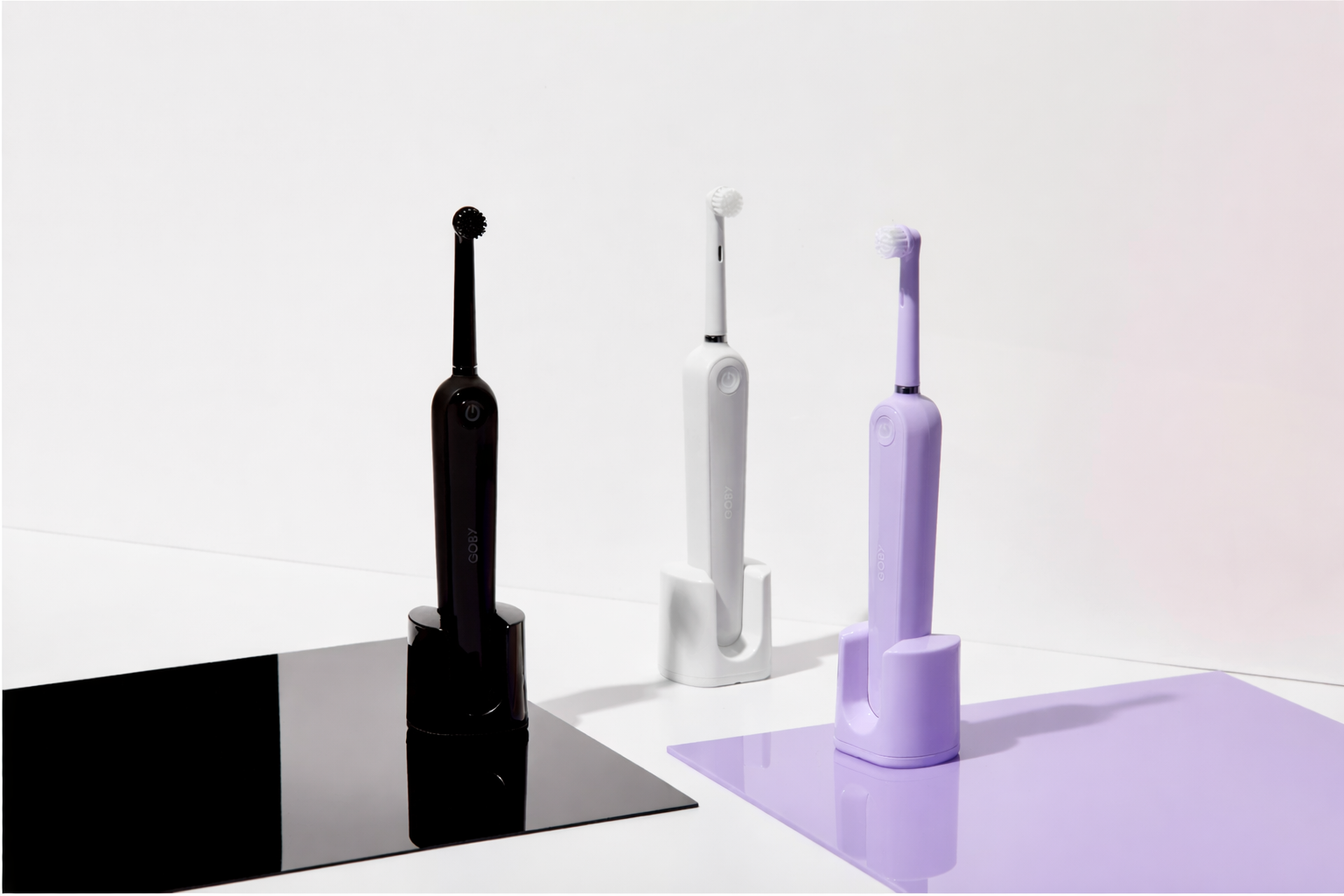 Three toothbrushes, one black, one white, and one purple, standing in their charging stations on a white surface with a plain white background.