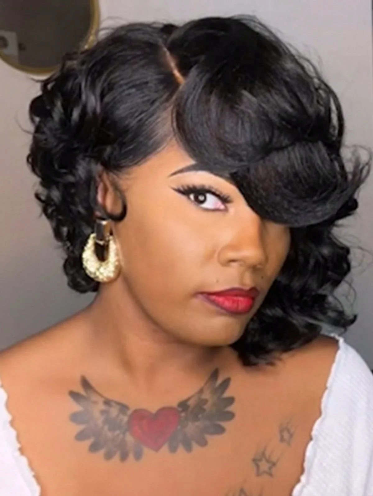 A woman with dark, curly hair styled in voluminous layers, wearing large gold hoop earrings with white stones, red lipstick, and a white top. She has tattoos on her chest including a red heart with wings and some smaller designs.