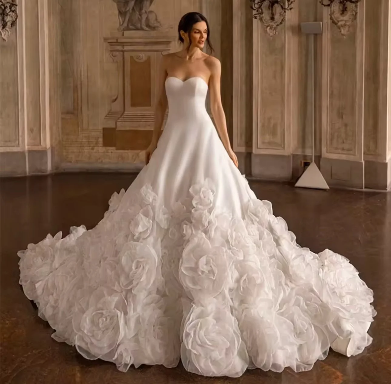 A woman in a strapless wedding gown with a fitted bodice and a full skirt made of large, textured fabric roses, standing in a decorated room with ornate wall panels.