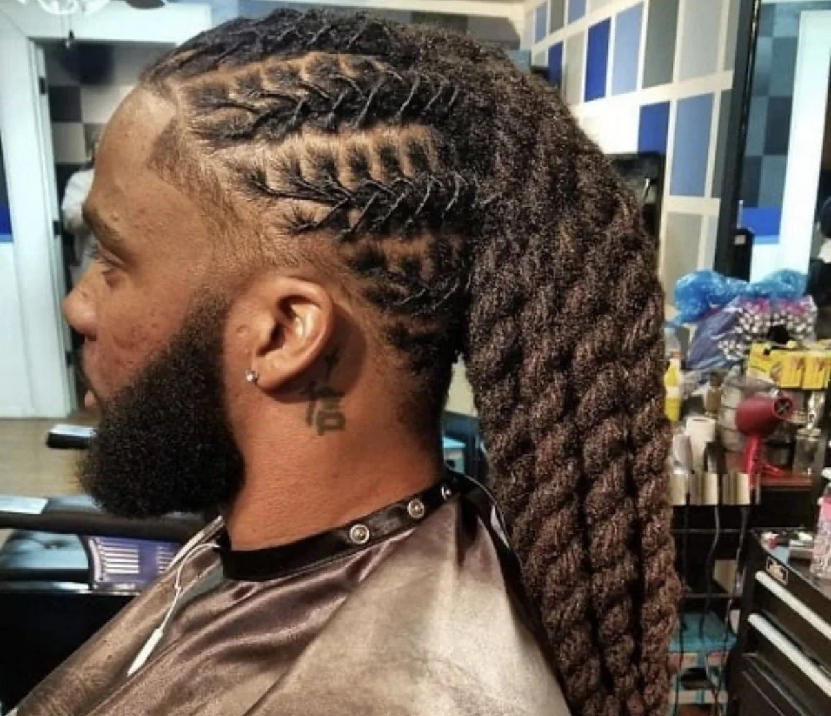 A man with braided and dreadlocked hair, a beard, and an earring is sitting in a salon chair.