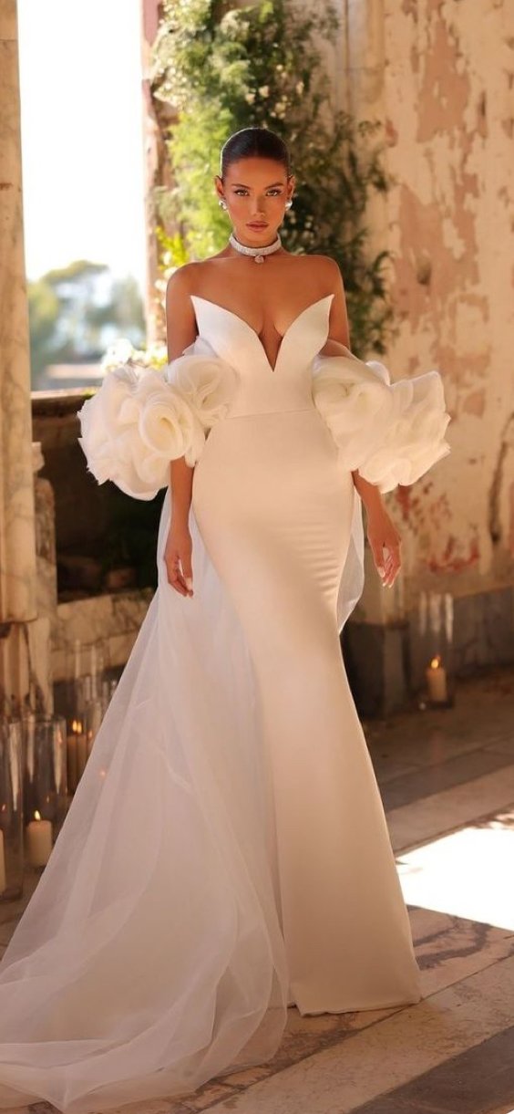 A woman in an elegant white wedding gown with large puffed sleeves and a deep V neckline standing indoors near a window with candles and greenery in the background.