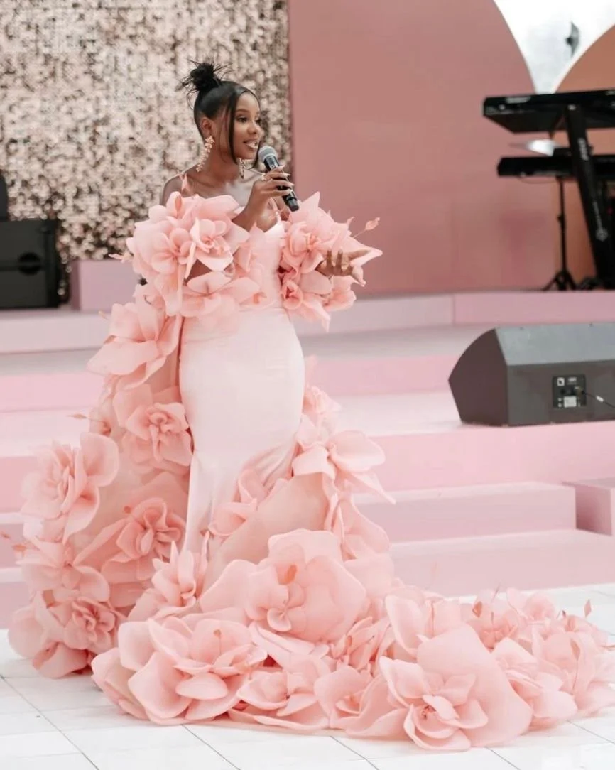 A woman in an elegant pink gown with floral embellishments is standing on a stage, holding a microphone, with musical equipment in the background.