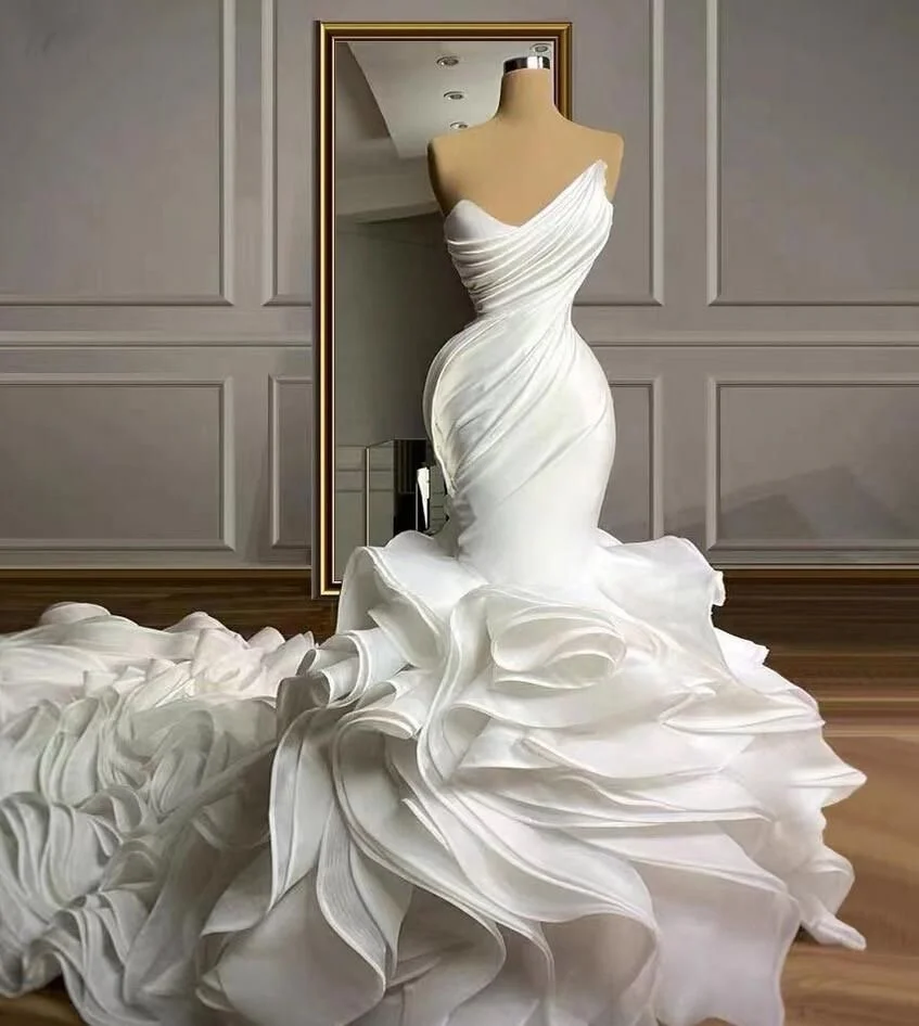 A white wedding dress on a mannequin seen in front of a mirror, with the dress's elaborate ruffled train flowing onto the floor, set against a wall with decorative paneling.