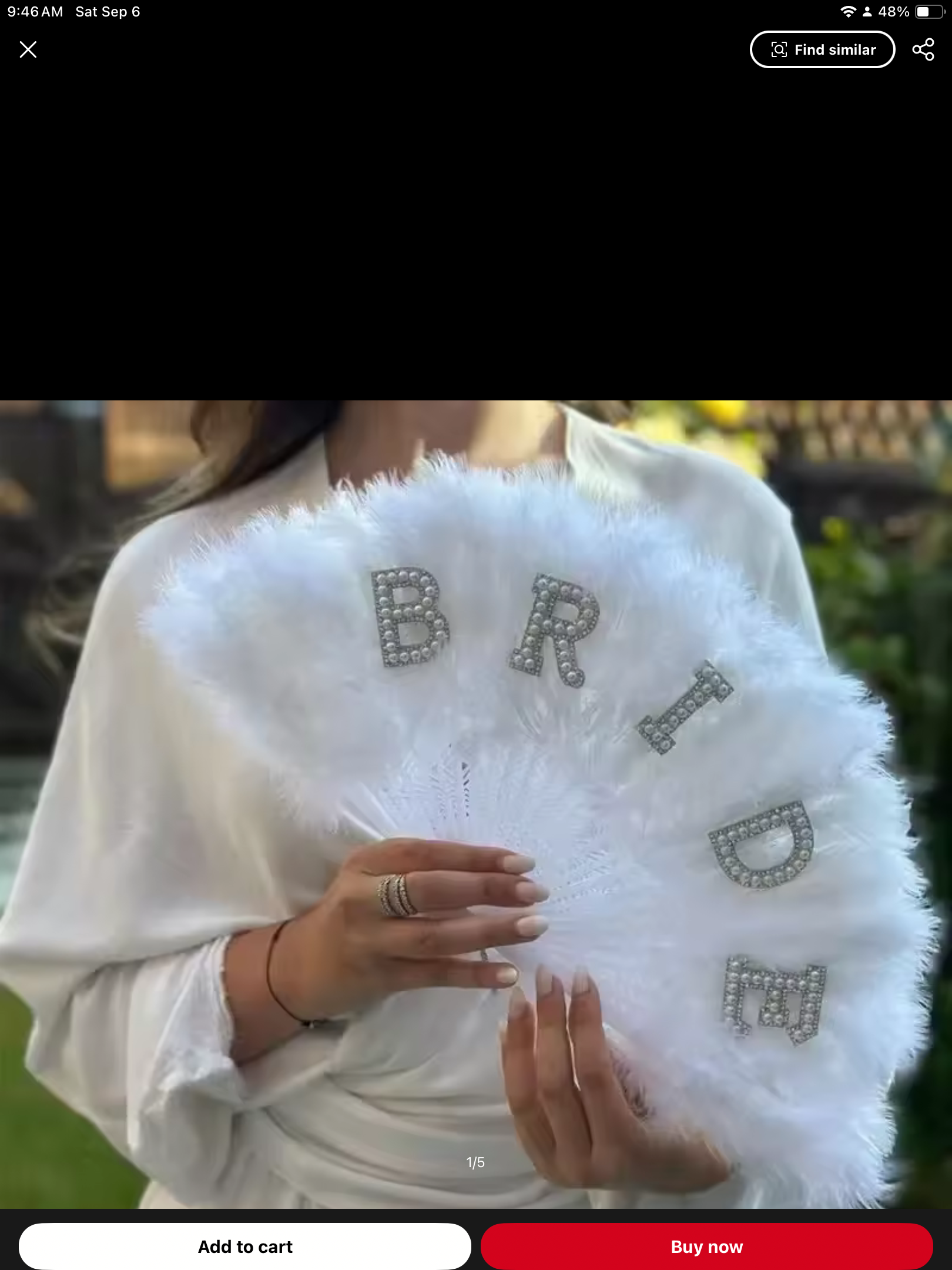 Woman holding a white feather fan with the word 'BRIDE' decorated with rhinestones. She is wearing a white dress and rings, standing outdoors with greenery in the background.