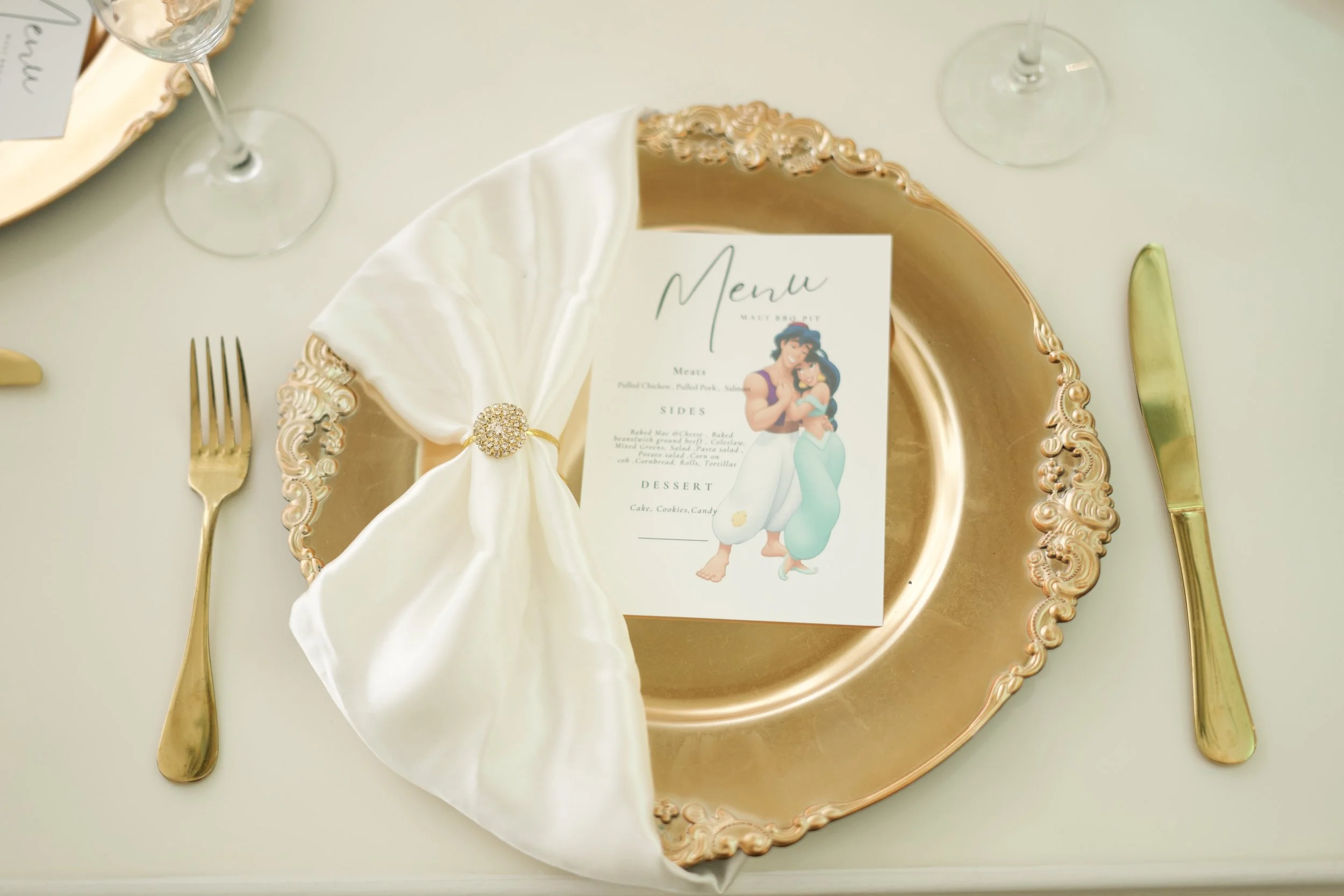 Elegant dining table setting with a gold ornate plate, a white napkin with a decorative brooch, gold cutlery, and a printed menu featuring a cartoon couple, the scene includes wine glasses and a cream-colored tablecloth.