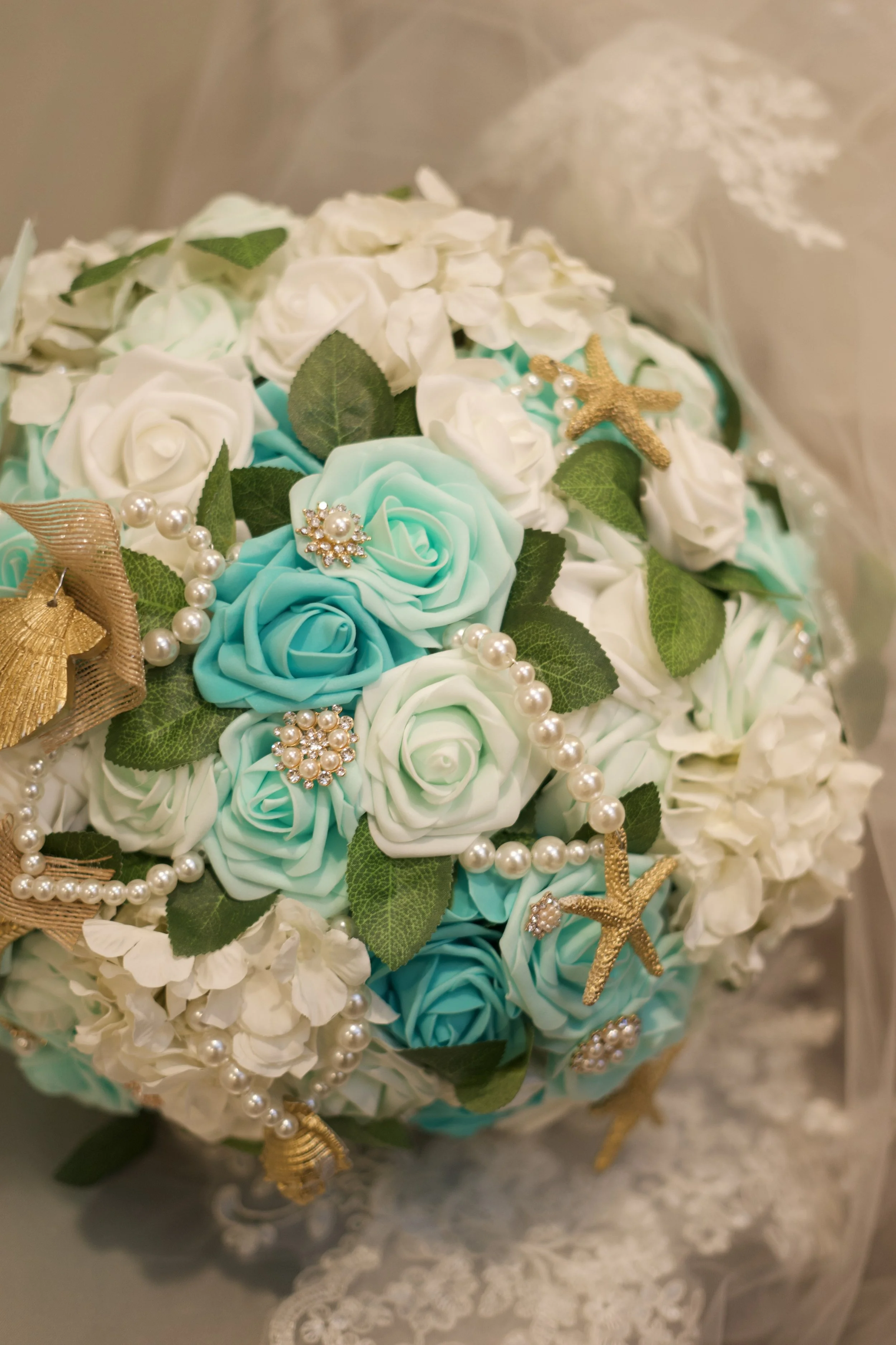 A bouquet with white and blue roses, green leaves, and decorative gold starfish, pearl strands, and pearl brooches.