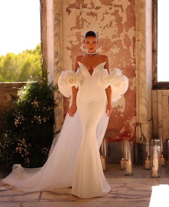 A woman in an elegant white wedding or evening gown with dramatic puffed sleeves and a fitted silhouette, standing indoors against a weathered rustic wall with large windows and candles on the floor.