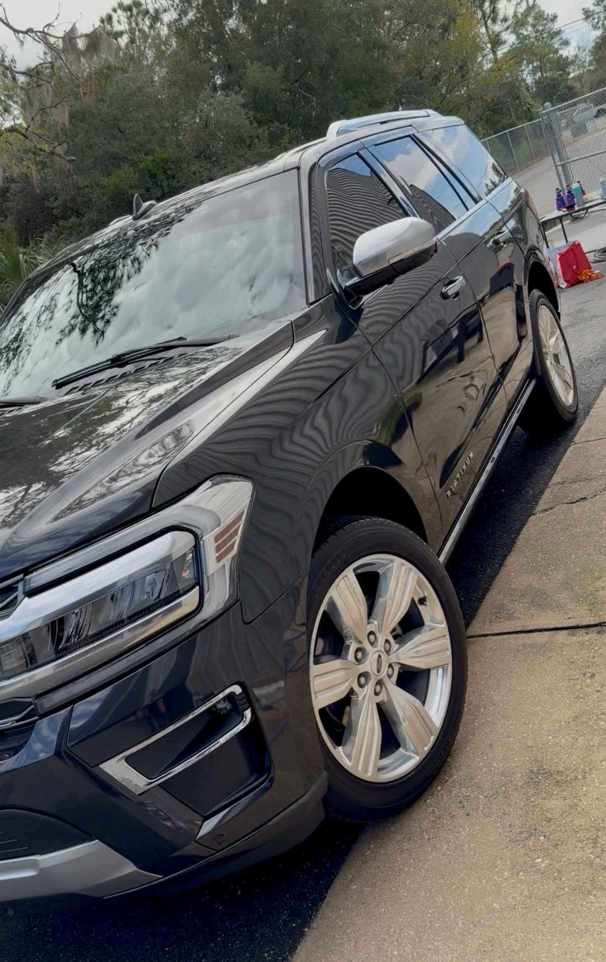 A black SUV parked on a driveway with trees in the background, some items on a table nearby, and a fenced outdoor area.