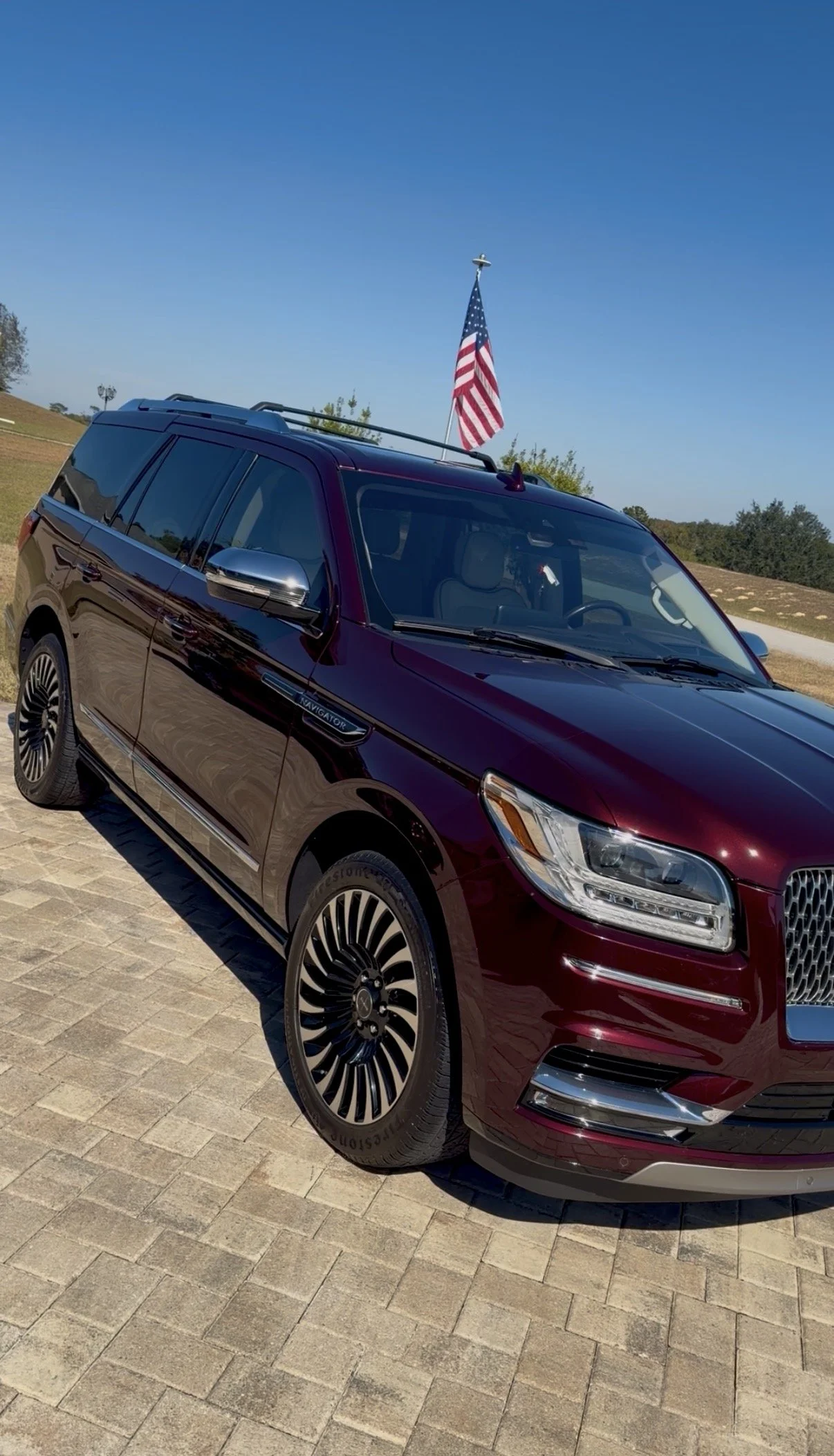 A maroon Mercedes-Benz SUV with an American flag mounted on the roof, parked on a brick driveway under a clear blue sky.