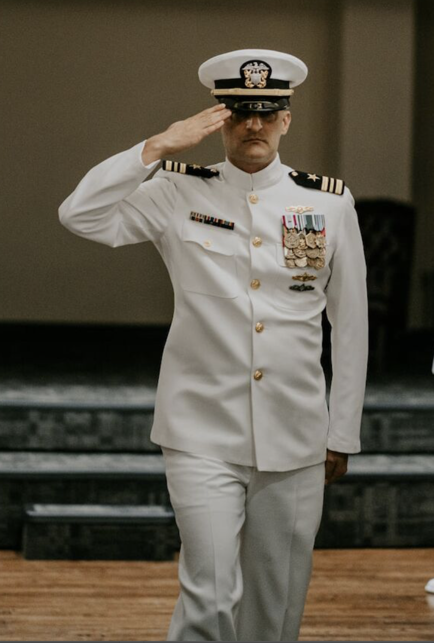 A man in a white military uniform salutes with his right hand, wearing a white peaked cap with a black visor and gold insignia, decorated with medals and ribbons.