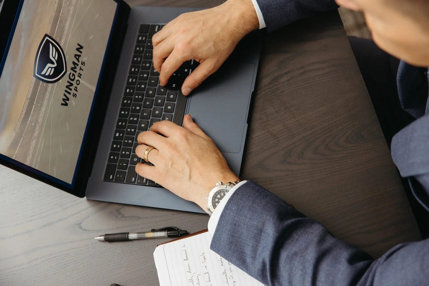 A person in a suit typing on a laptop with a 'WINGMAN SPORTS' logo on the screen, on a dark wooden table, with a notebook and pen nearby.
