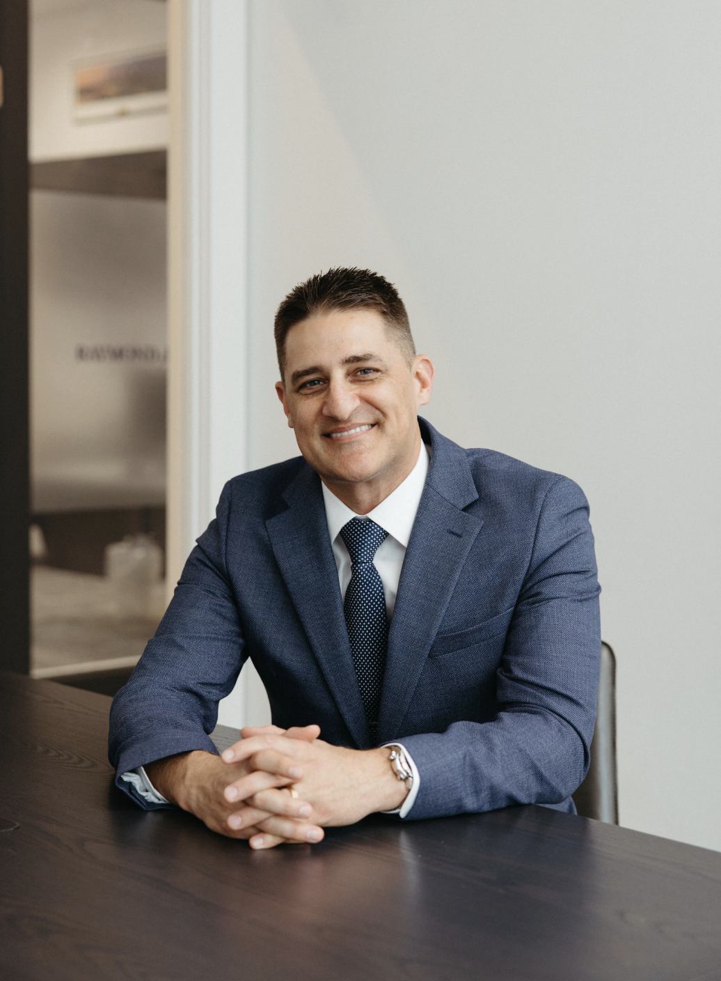 A smiling man in a blue suit with a white shirt and a navy polka dot tie, sitting at a dark wooden table with hands clasped, in a professional office setting.