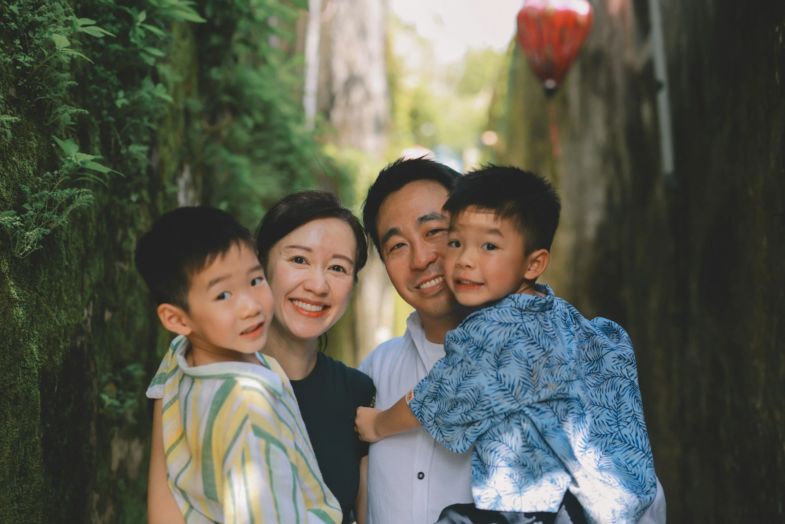 A happy family of four with two young boys, hugging and smiling at the camera outdoors between moss-covered rocks with greenery and red lanterns hanging in the background.