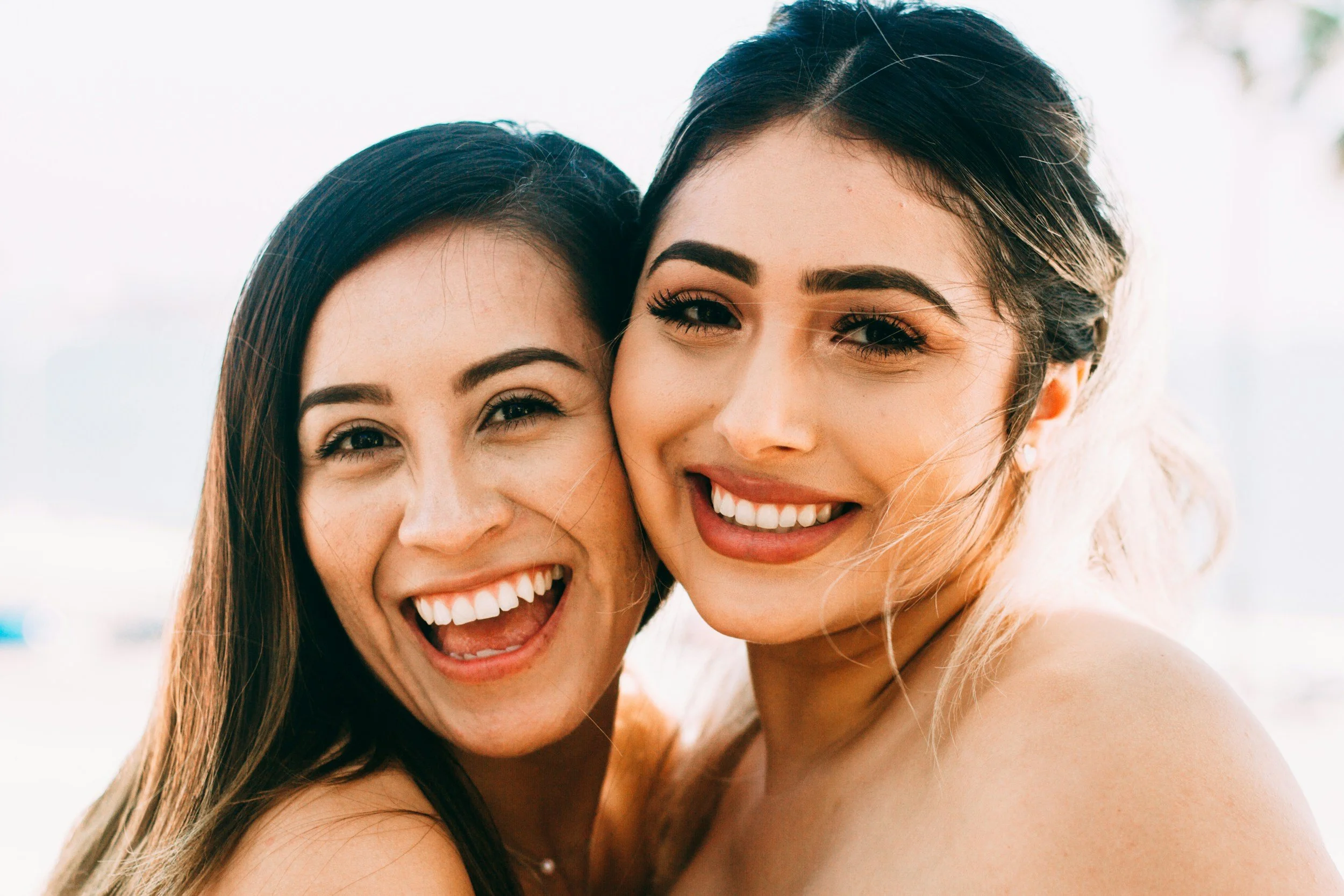 Close-up of two smiling women with dark and light hair, outdoors on a sunny day.