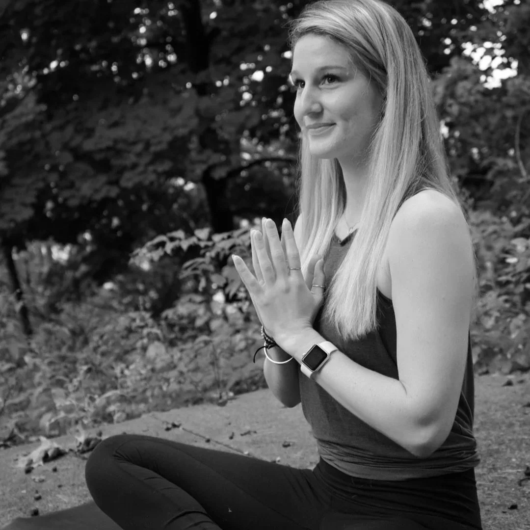 A young woman practicing yoga outdoors, sitting cross-legged with hands in prayer position, smiling gently, surrounded by trees and nature.