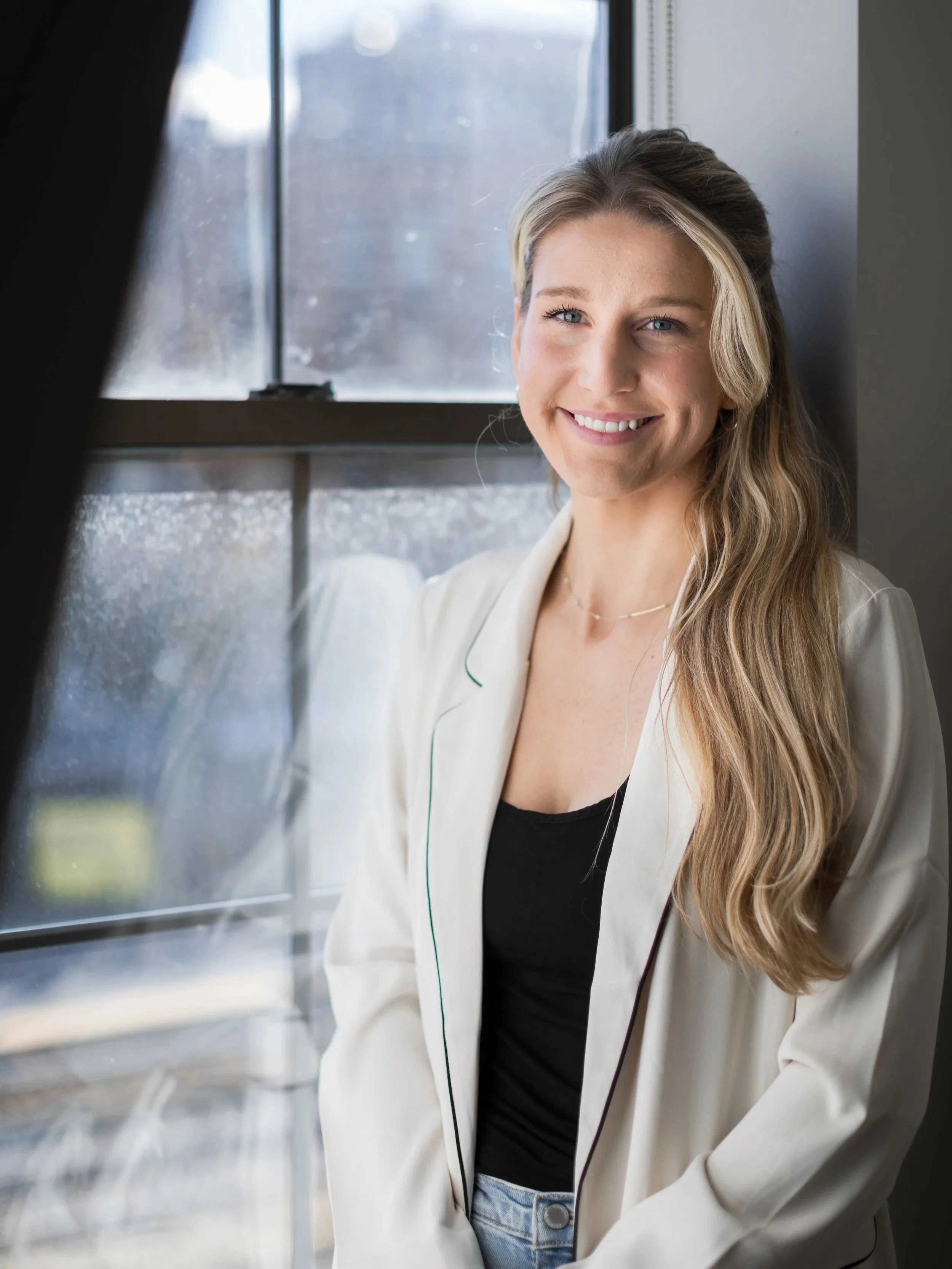 A young woman with long blonde hair smiling, standing by a window with natural light.