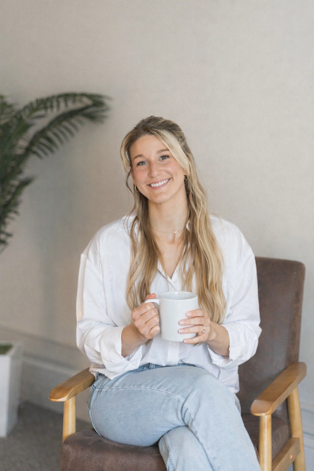 A woman with long blonde hair smiling and holding a white mug, sitting in a wooden chair in a cozy, neutral-toned room.