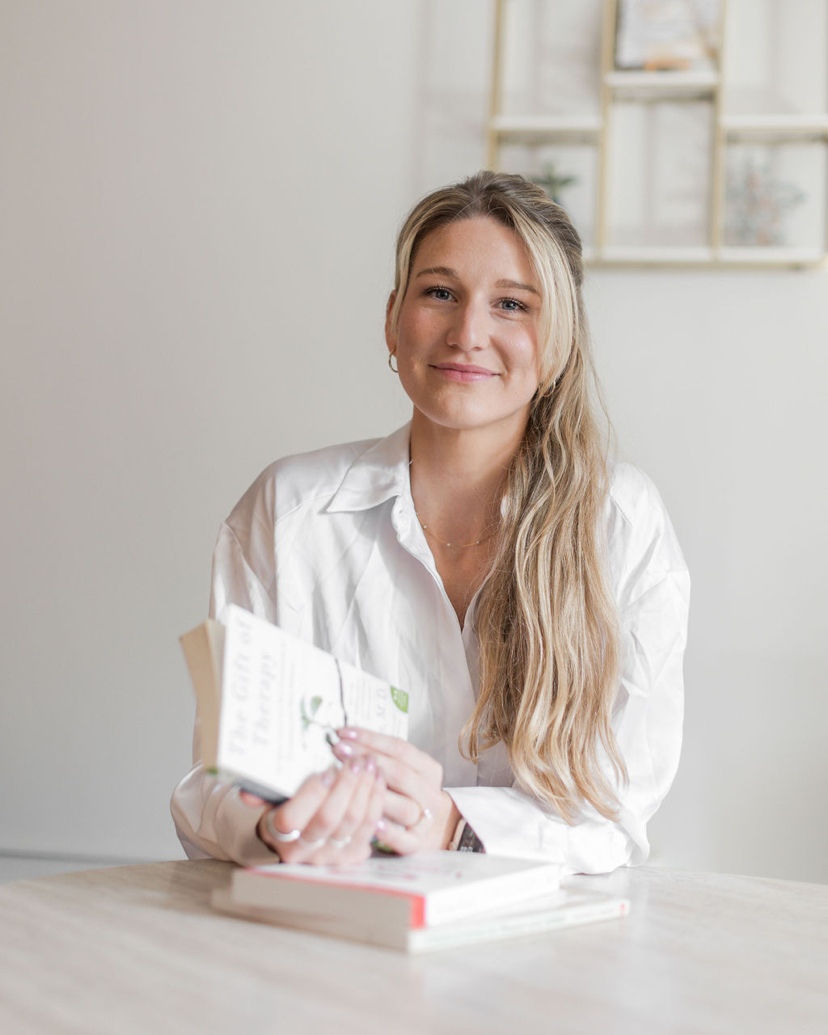 A young woman with long blonde hair, wearing a white shirt, sitting at a table with a book in her hands and a closed book in front of her. She is smiling and looking at the camera, with a blurred shelf in the background.