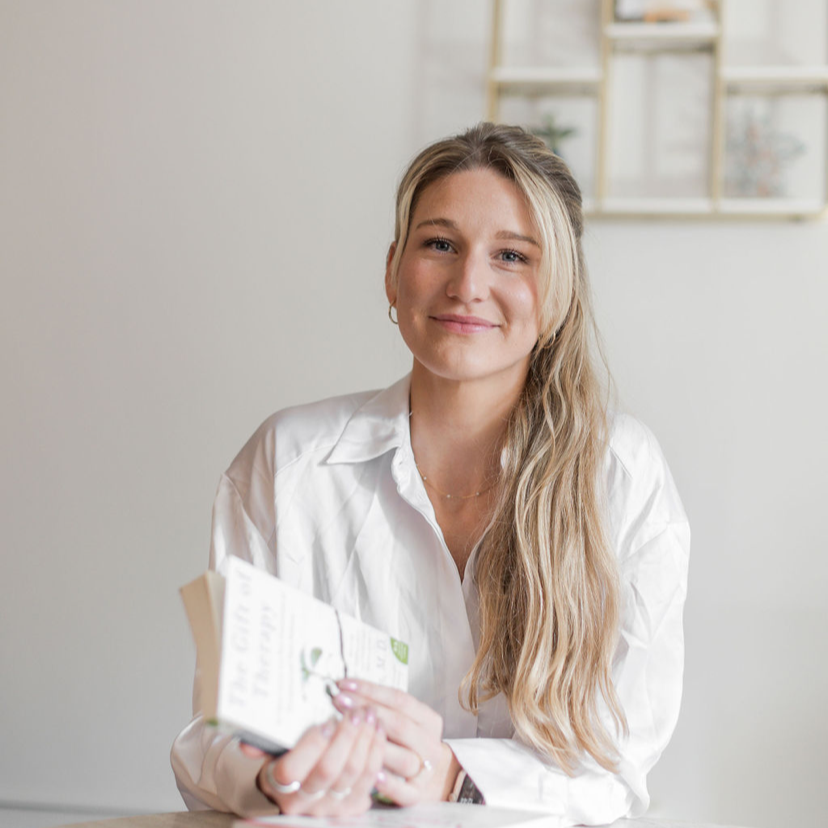 A young woman with long blonde hair and a white shirt sitting at a table, holding a pamphlet or booklet, smiling in a well-lit room with bookshelf on the background.