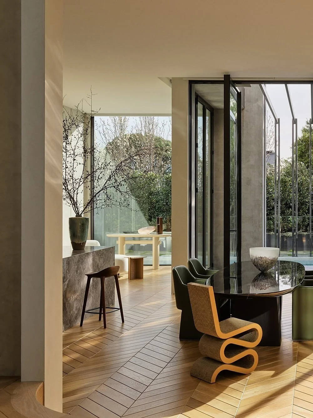 Intérieur moderne avec grand espace de salle à manger, cheminée en pierre, chaises différentes autour d'une grande table en verre, et une vue sur le jardin avec des arbres à travers de grandes fenêtres.