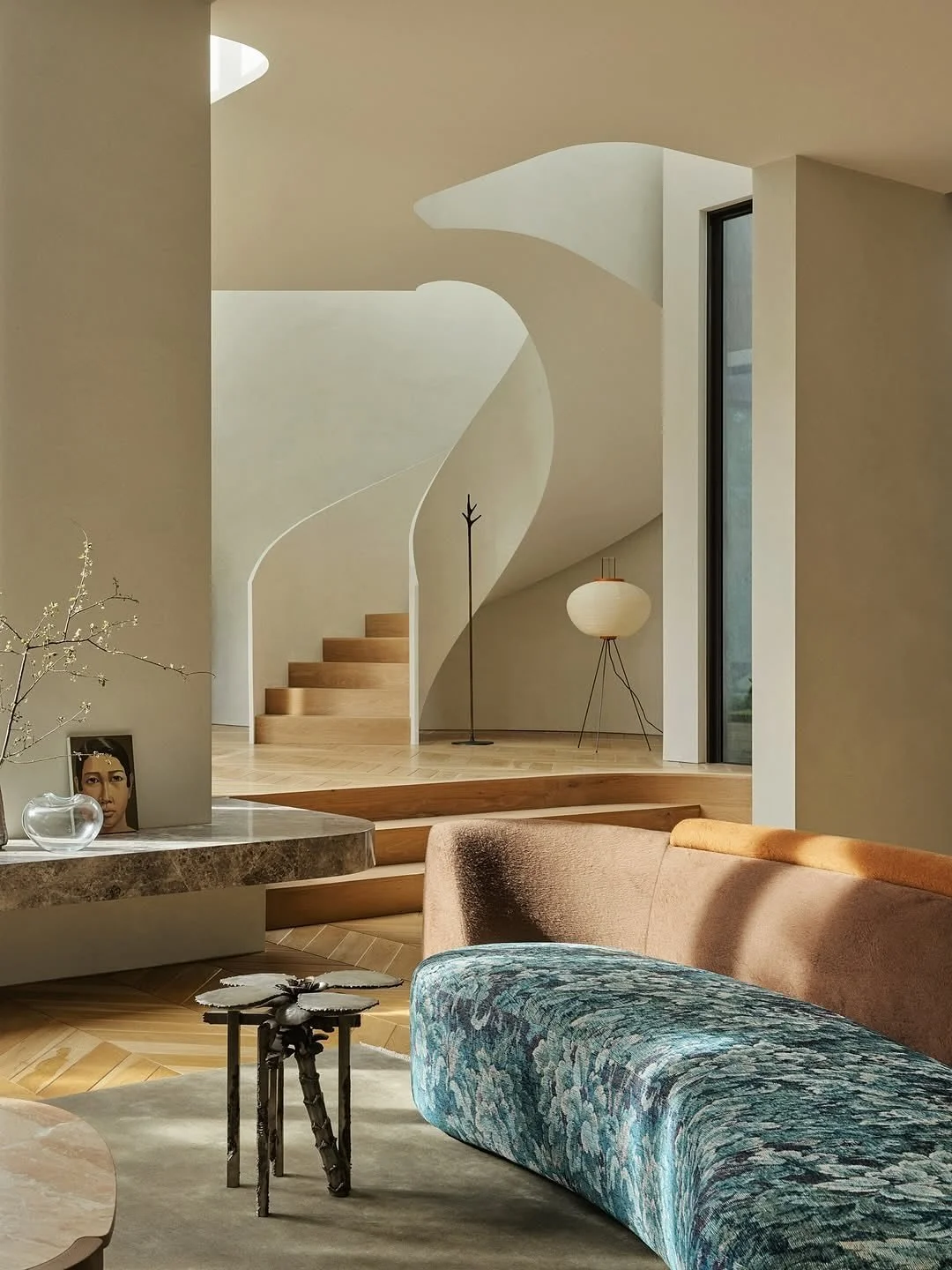 Intérieur moderne avec canapé beige et fauteuil en tissu coloré, table basse artistique, mur blanc avec escaliers en bois, lampadaire en papier et grande fenêtre.