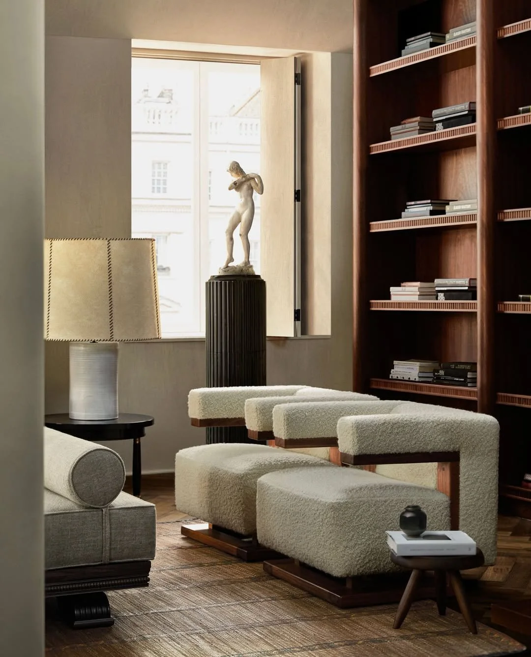 Salon avec fauteuils en cuir beige, lampe à abat-jour, sculpture classique près de la fenêtre, bibliothèque en bois, et petit meuble avec une vase noire et des livres.