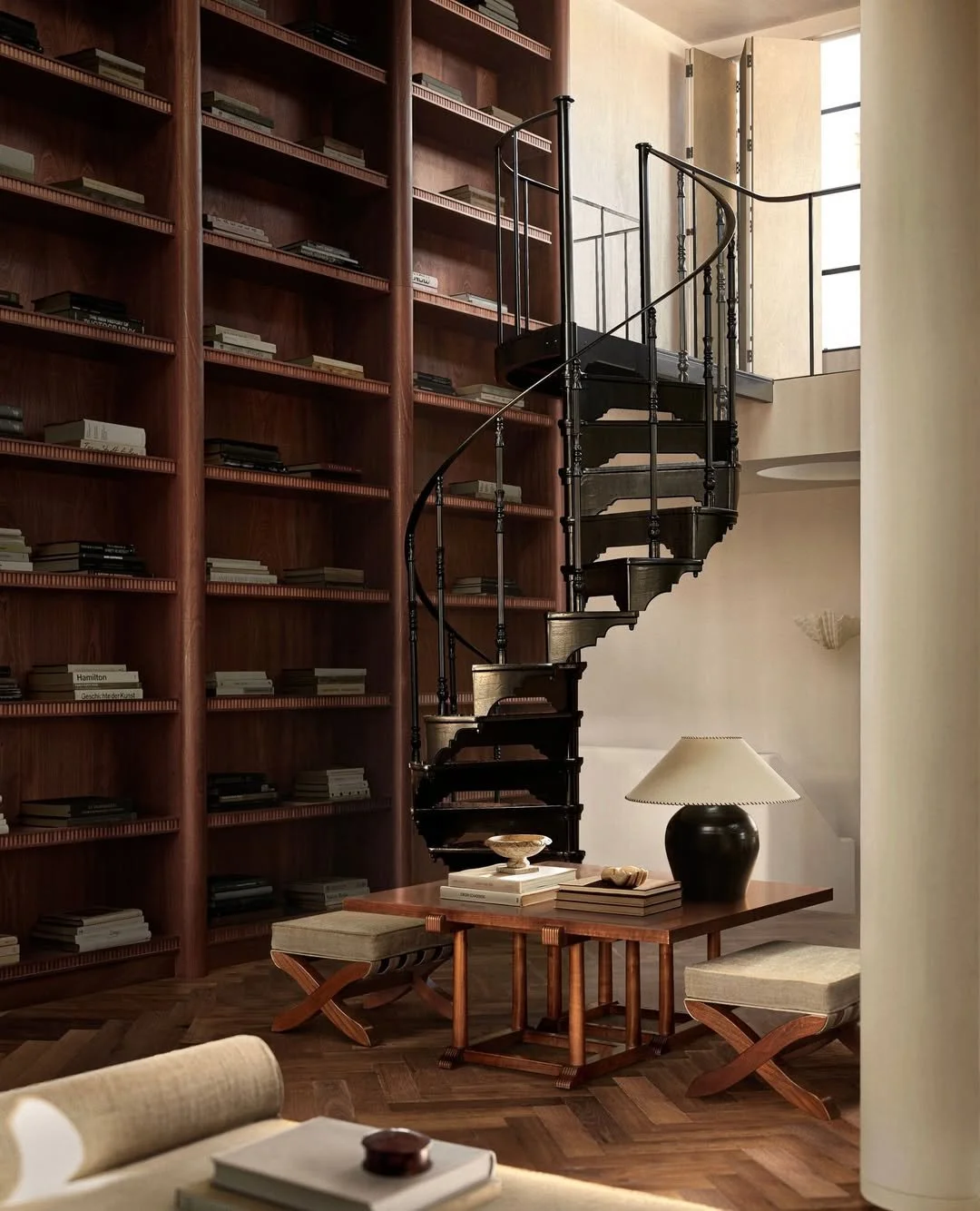 Intérieur d'une pièce avec une grande bibliothèque en bois, une table en bois avec une lampe, des livres, et des sièges. Un escalier hélicoïdal noir est visible.