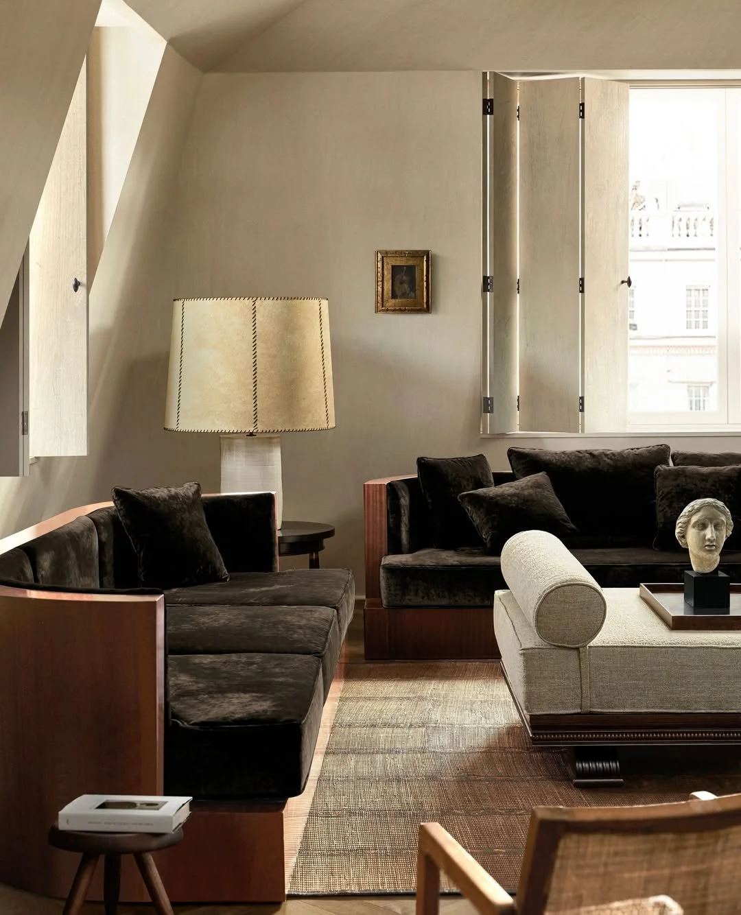 Salon avec deux canapés en velours foncé, une lampe beige, un tableau encadré, un bureau blanc avec une sculpture de visage, une table basse en bois avec un rouleau décoratif, une fenêtre ouverte et un tapis beige et marron.