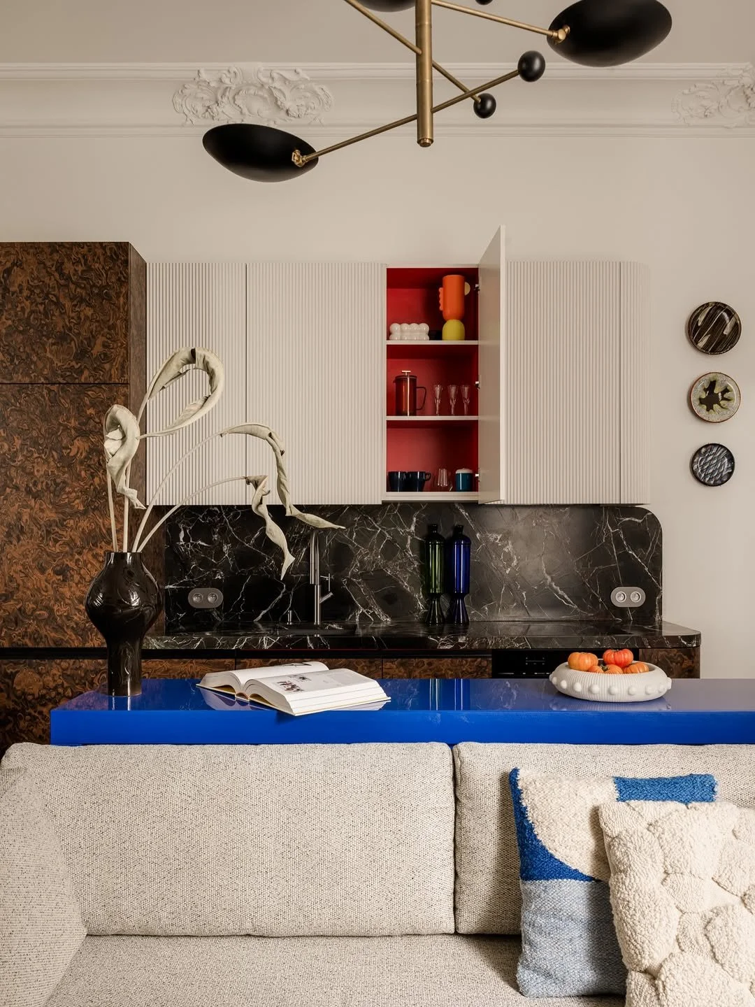 Cuisine moderne avec un comptoir en marbre noir, armoires blanches et en bois, étagères ouvertes avec vaisselle colorée, chandelier design, canapé beige avec coussins, vase avec des branches sèches, livre ouvert et plat de tomates sur la table bleue.