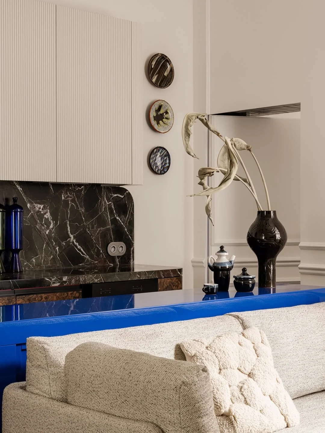 Intérieur moderne avec un canapé beige, une table avec des vases noirs, un vase marron avec des branches séchées, et un mur avec trois petits tableaux décoratifs.