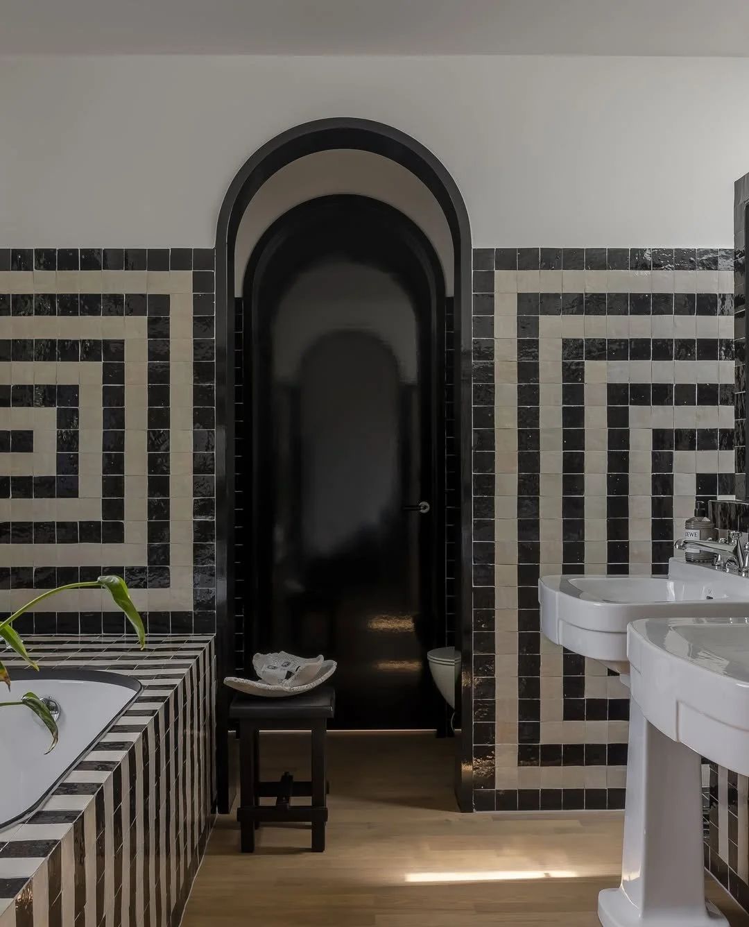 Salle de bain avec une mosaïque noire et blanche, deux lavabos, un miroir, un placard noir, et un tapis avec serviette sur un banc.