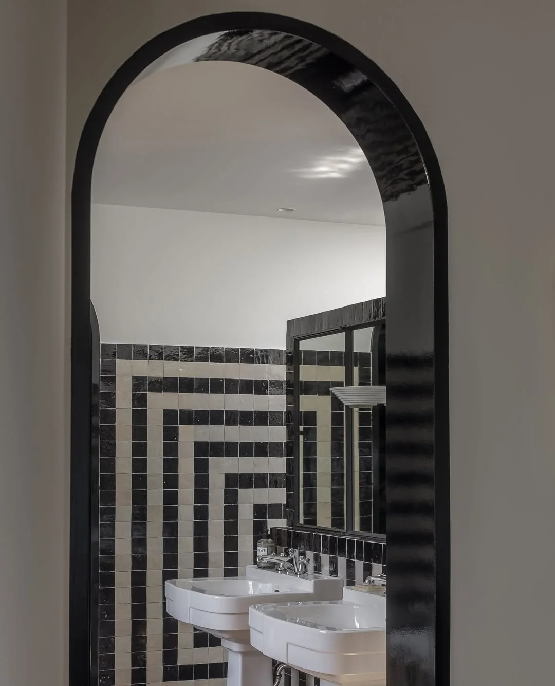 Vue d'une salle de bain à travers une arche avec miroir et lavabos doubles, murs en carrelage noir et beige avec motif géométrique.