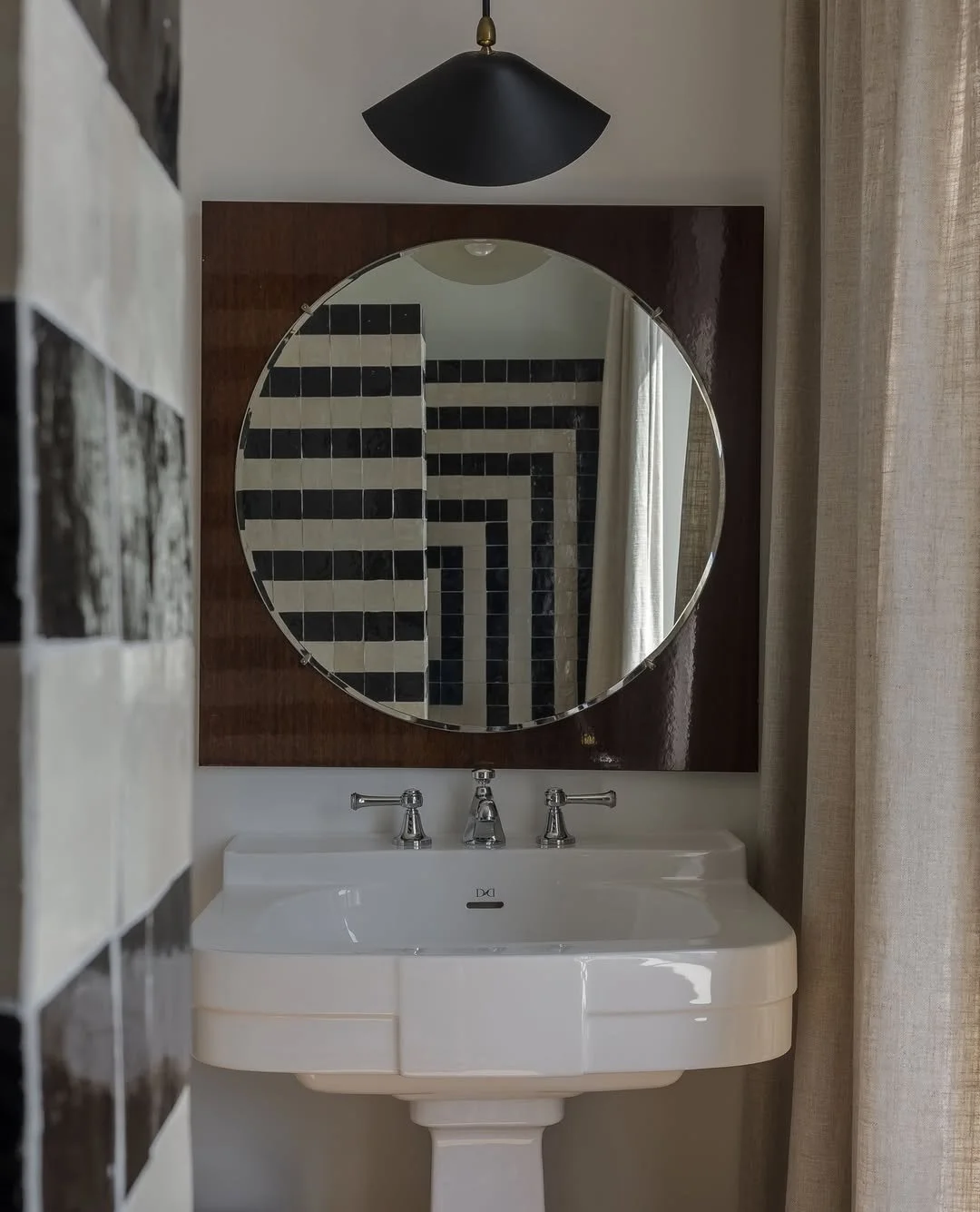 Miroir rond au-dessus d'un lavabo blanc avec deux robinets en chrome, dans une salle de bain avec un panneau en bois derrière le miroir, et des carreaux noirs et blancs en arrière-plan.