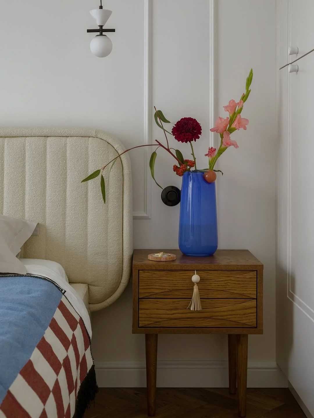 Une table de nuit en bois avec une grande vase bleue contenant des fleurs roses et rouges, à côté d'un lit avec une tête de lit beige et une couverture à motifs rouges et blancs, dans une chambre à décor minimaliste.