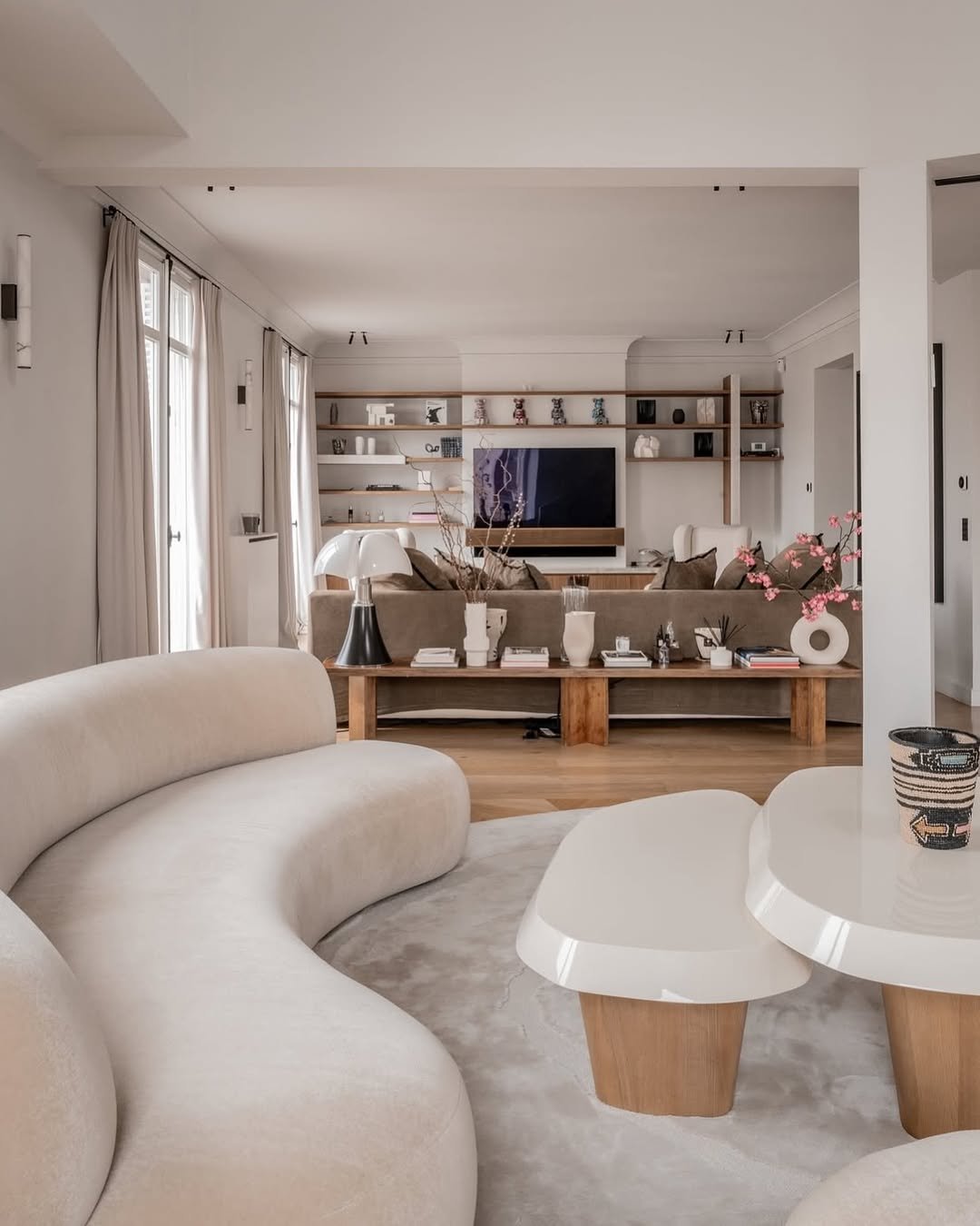 Intérieur d'un salon moderne avec canapé beige, table basse en bois et décoration élégante, vue vers un mur avec étagères et télévision.