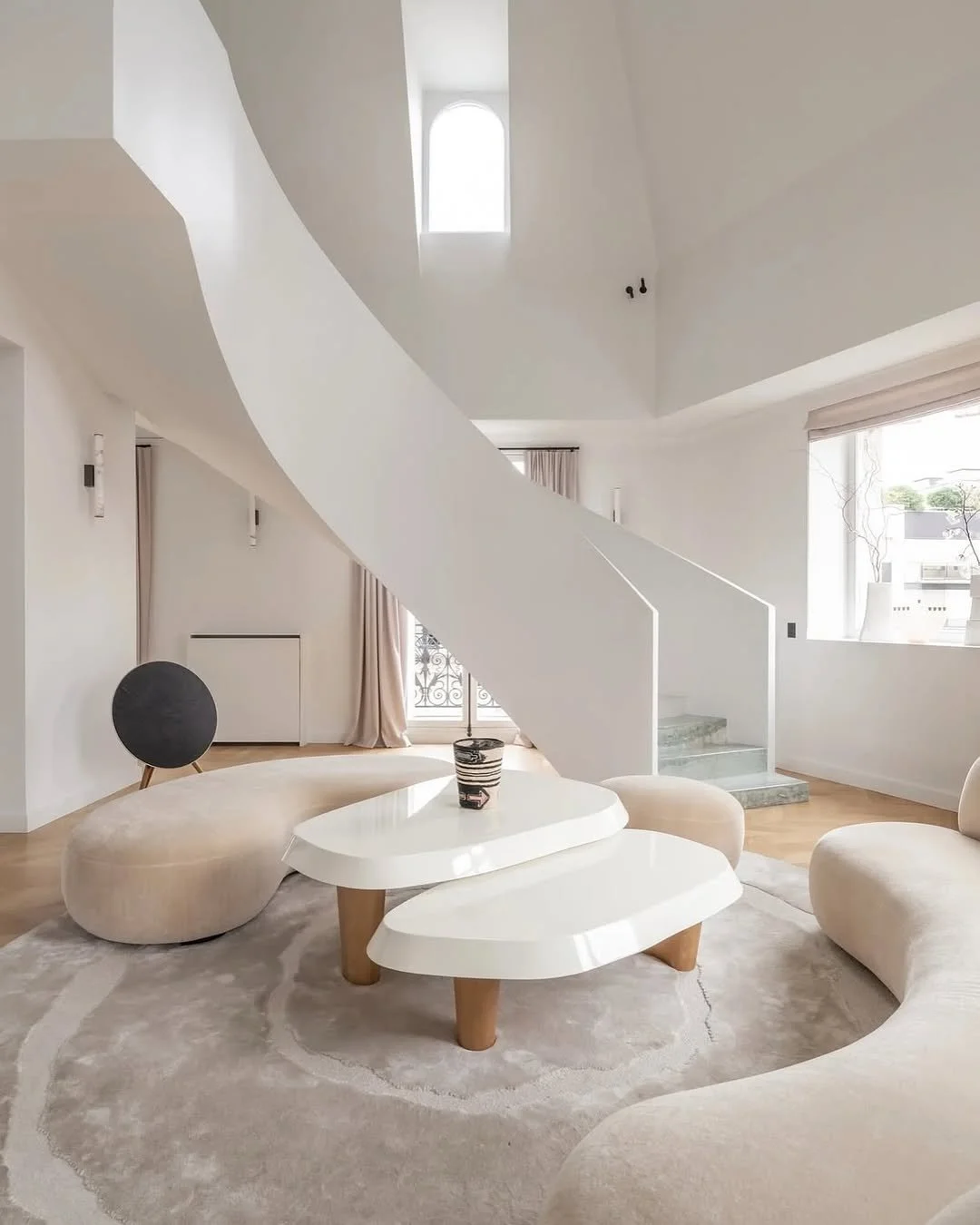 Salon moderne avec canapé beige, table basse blanche, escalier blanc en colimaçon et grande fenêtre laissant entrer la lumière naturelle.